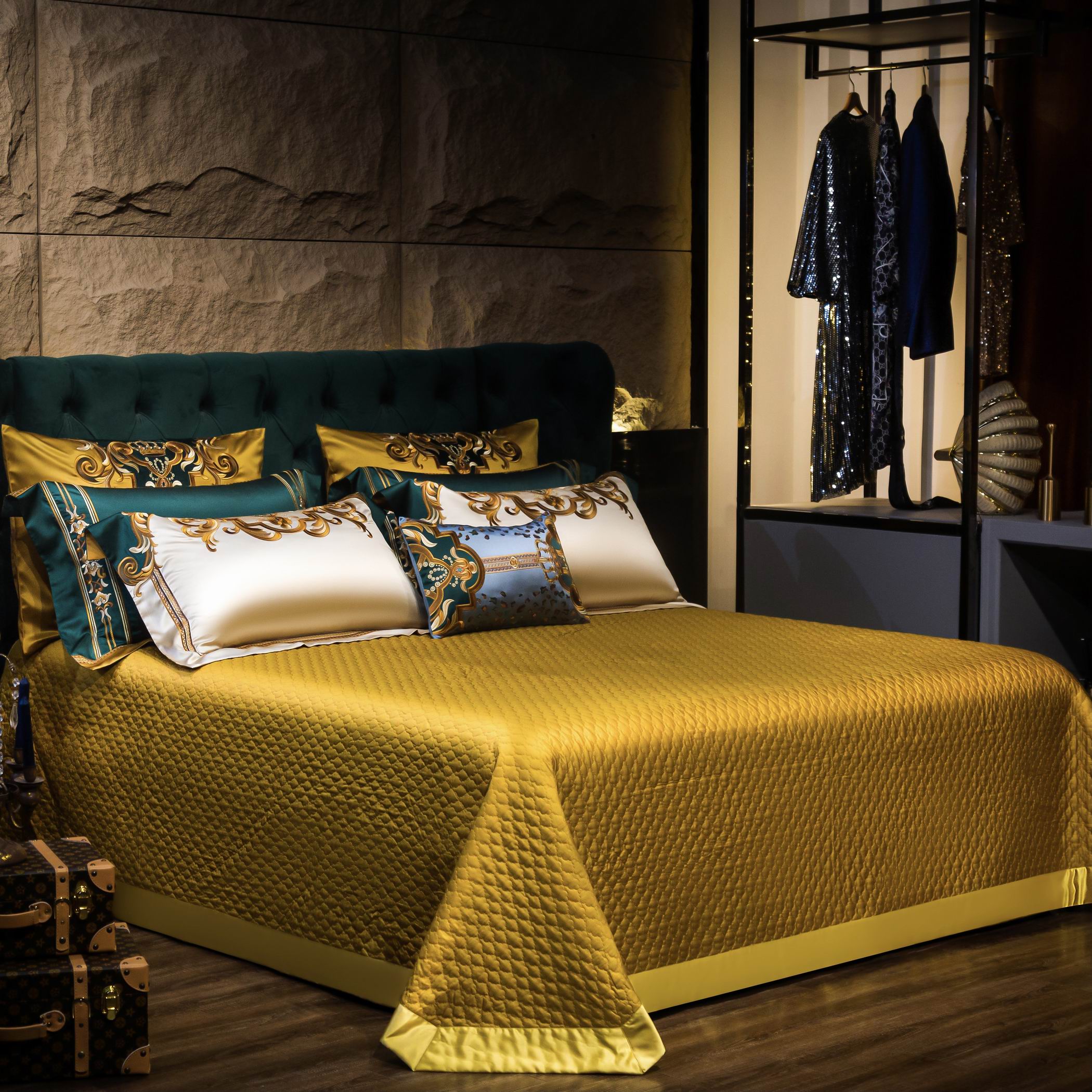 Luxurious bedding display featuring a rich gold quilted bedspread paired with emerald green and cream pillows adorned with intricate baroque embroidery. The elegant setting is complemented by a plush emerald green headboard and a chic wardrobe in the background showcasing sparkling attire.