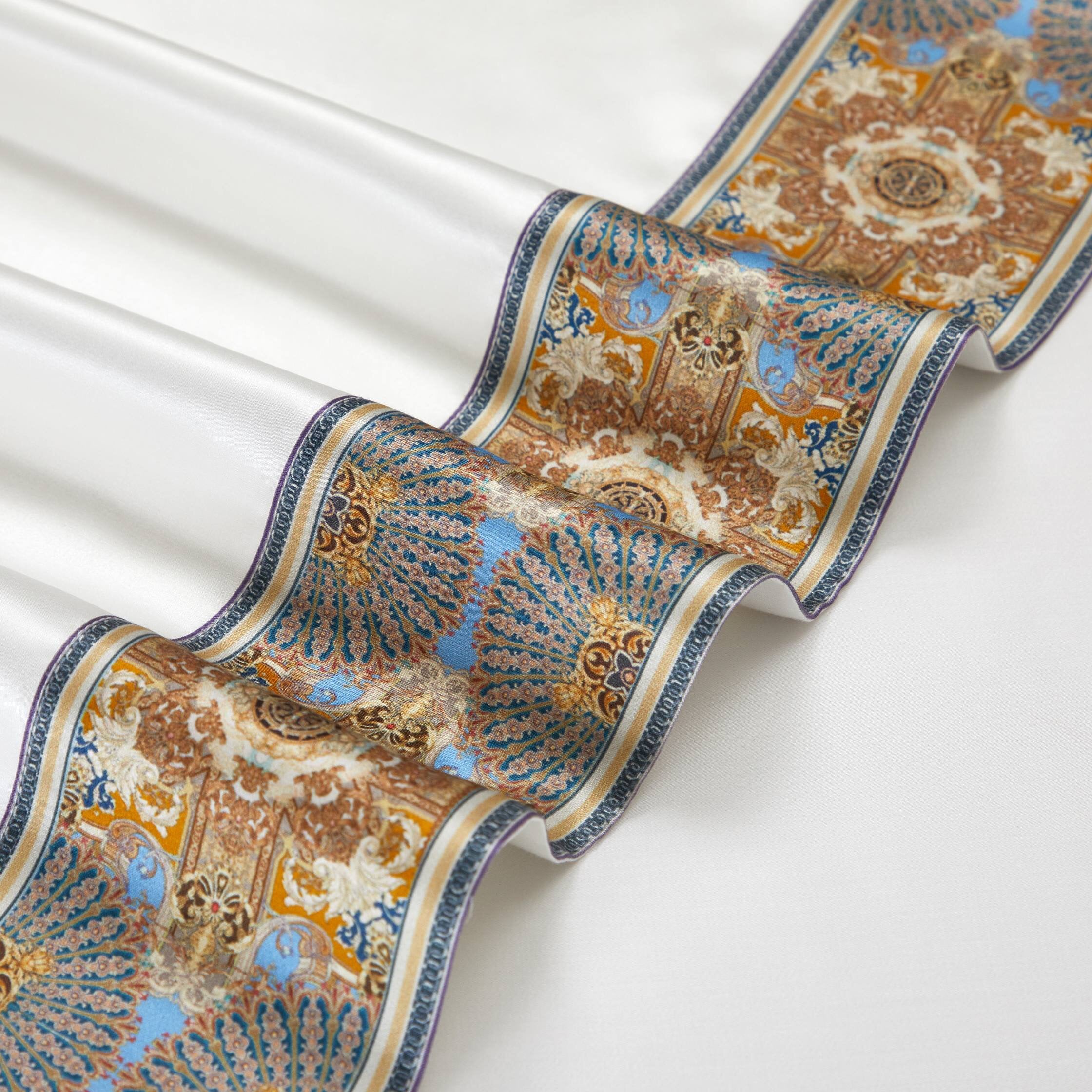 Close-up of a luxurious fabric featuring a silky white background adorned with an intricate border design. The border showcases ornate golden and blue motifs with detailed patterns, creating a regal and sophisticated appearance.