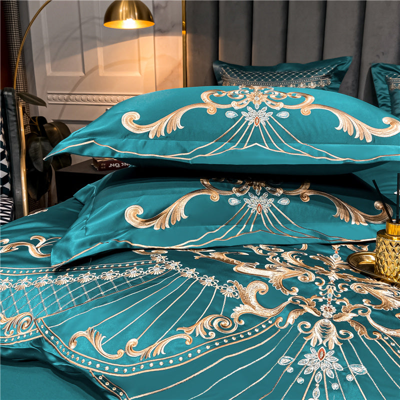 Green bedding set with intricate gold embroidery featuring swirling floral patterns and symmetrical designs. Stacked pillowcases and the duvet cover display ornate detailing with fine stitching. A gold diffuser and tray are positioned on the side, enhancing the luxurious ambiance.

