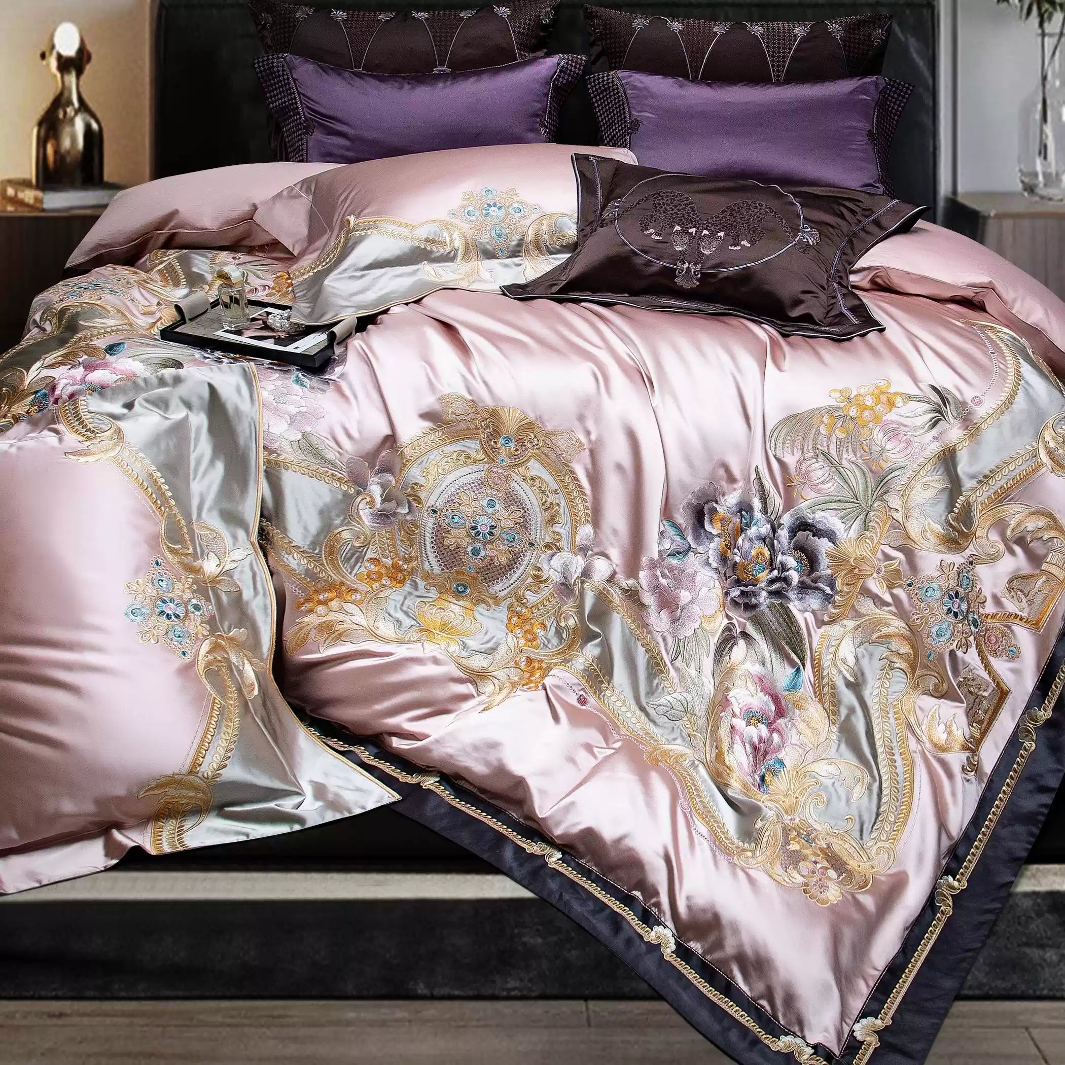 An exquisite close-up of a luxurious blush pink bedding set adorned with elaborate gold embroidery and delicate floral patterns. The silk-like fabric features intricate details, including shimmering accents of lavender and pastel tones, exuding elegance and sophistication. Deep plum and lavender pillows in the background add a complementary contrast, enhancing the set's opulent appearance.