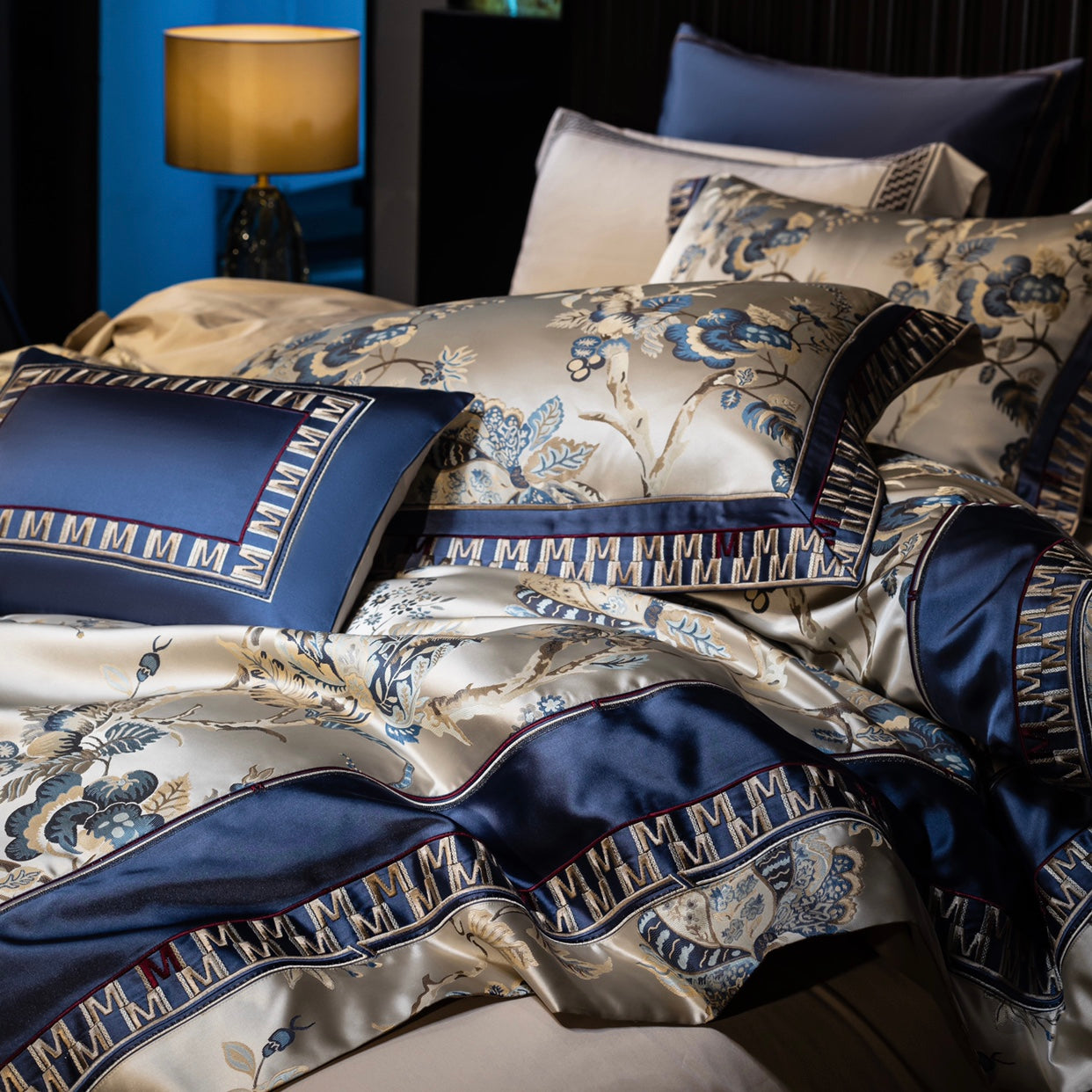 A close-up of an opulent bedding set showcasing a champagne-toned fabric adorned with intricate blue floral embroidery and elegant patterns. The design is framed by rich navy blue satin borders, detailed with a distinctive "M" motif in geometric accents, adding a regal touch. A coordinating navy blue cushion complements the bedding, emphasizing its luxurious appeal. The combination of the glossy satin finish and embroidered details highlights the craftsmanship, making it a standout piece for a sophisticated