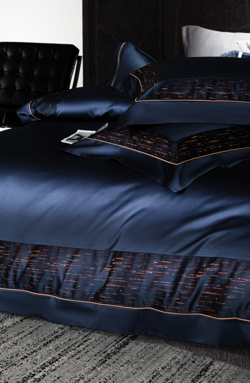 Navy blue bedding set featuring satin pillowcases with a glossy finish and a black border accented by orange and red streak patterns. The duvet cover showcases a matching border, creating a sleek and contemporary look. The bed rests on a textured gray carpet with a black leather chair in the background.

