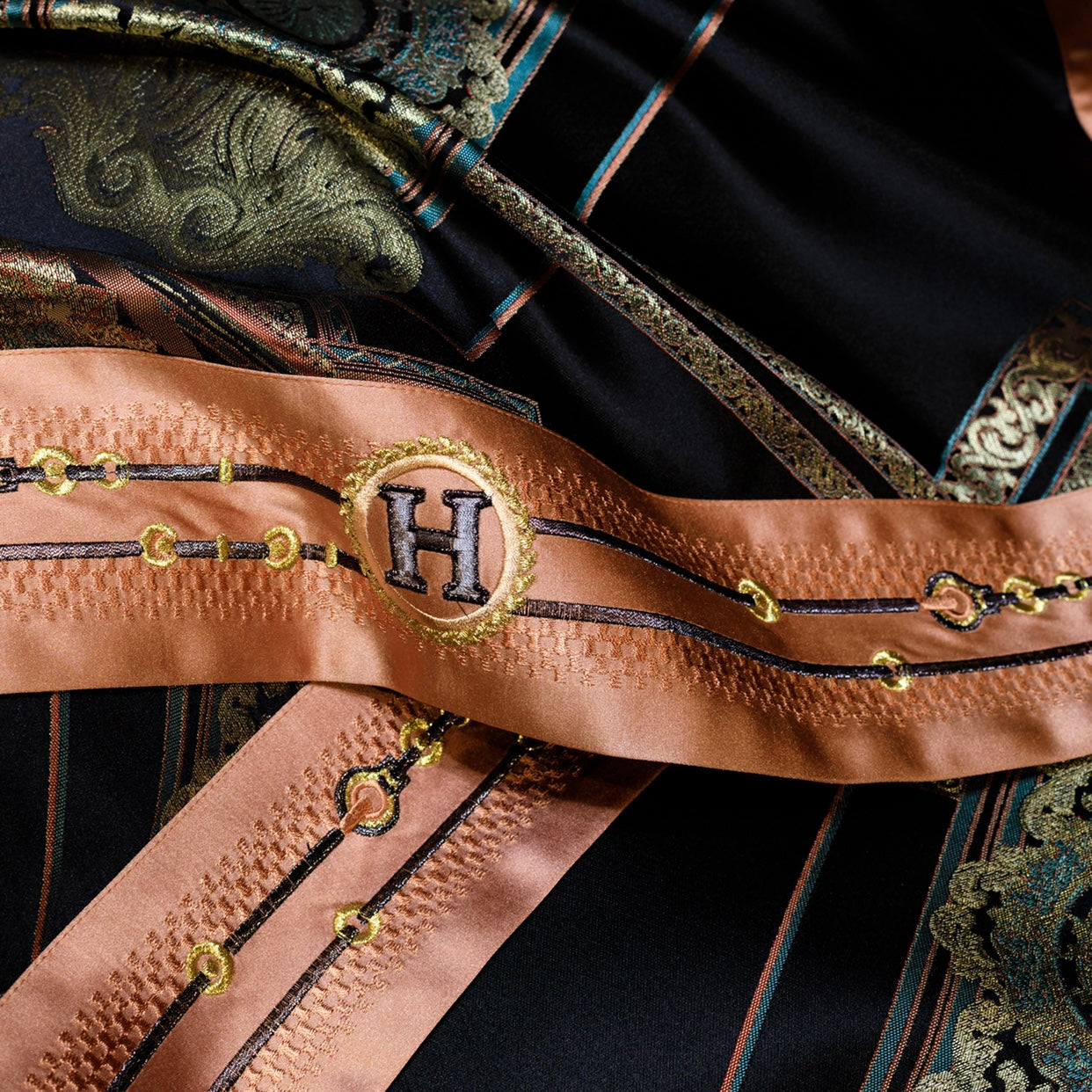 A close-up of luxurious bedding fabric showcasing a black base with intricate gold and copper embroidery. The highlight is a satin ribbon accent in a rich copper tone, adorned with elegant gold and black chain-like embroidery, and a prominent "H" monogram encircled by a gold wreath design. The detailed craftsmanship and metallic accents exude sophistication, adding a touch of opulence to the bedding's overall aesthetic.