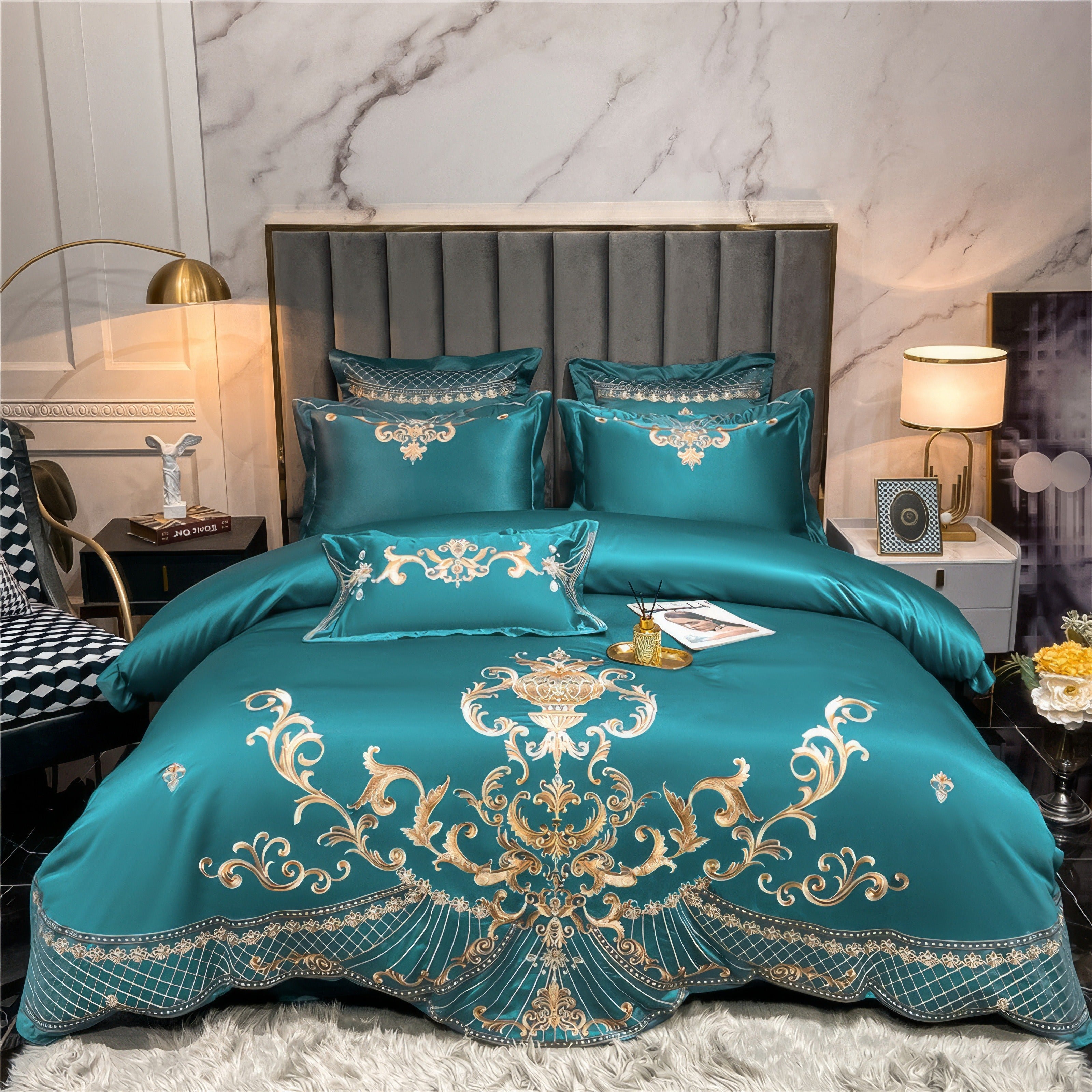 A teal satin bedding set with intricate gold embroidery featuring ornate scrollwork and floral details. The set includes matching pillows and a decorative lumbar pillow arranged on a neatly made bed. A plush white shag rug is spread at the foot of the bed, and soft lighting from a table lamp adds a cozy atmosphere. The elegant gray upholstered headboard and marble accent wall enhance the luxurious look of the bedroom.

