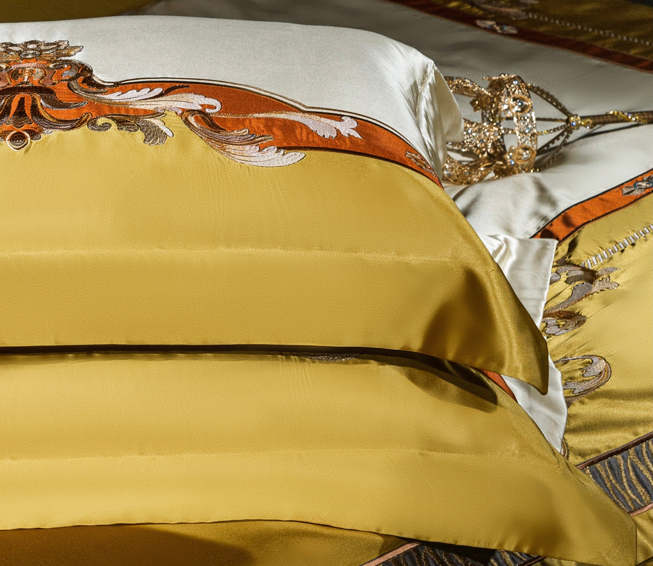 A close-up of luxurious bedding with golden satin pillowcases, highlighted by ornate baroque embroidery in vibrant orange and ivory tones. The intricate design adds a touch of regal sophistication, complemented by the smooth, lustrous texture of the fabric. The corner of a crown-shaped decor piece enhances the luxurious and opulent aesthetic of the setting.