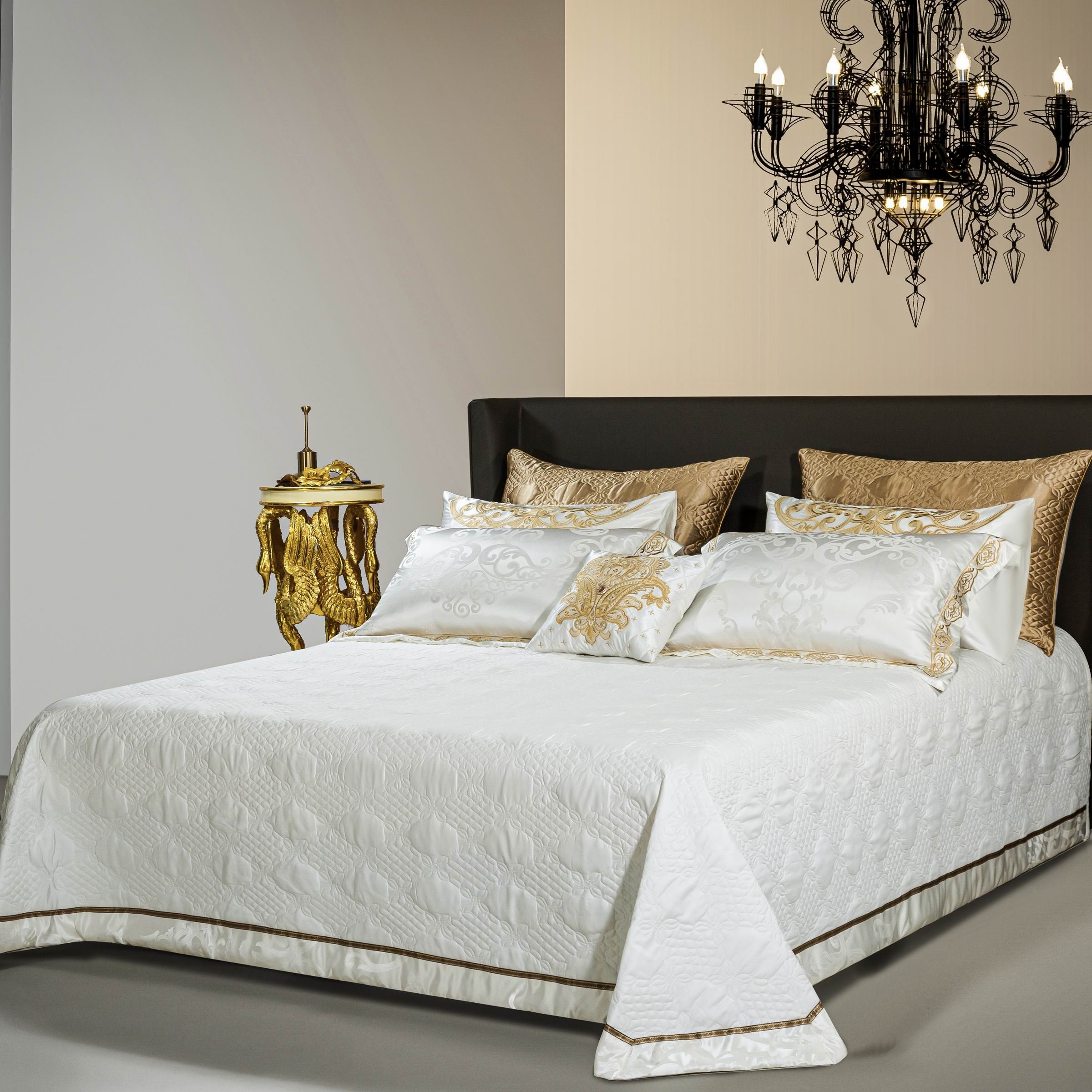 A luxurious bedroom setting featuring a white quilted bedding set adorned with subtle patterns and a touch of gold accents along the edges. The pillows showcase intricate gold embroidery, perfectly complementing the textured golden decorative pillows placed behind them. The ensemble is completed by a sophisticated black chandelier and an ornate gold side table, creating an opulent and harmonious ambiance.

