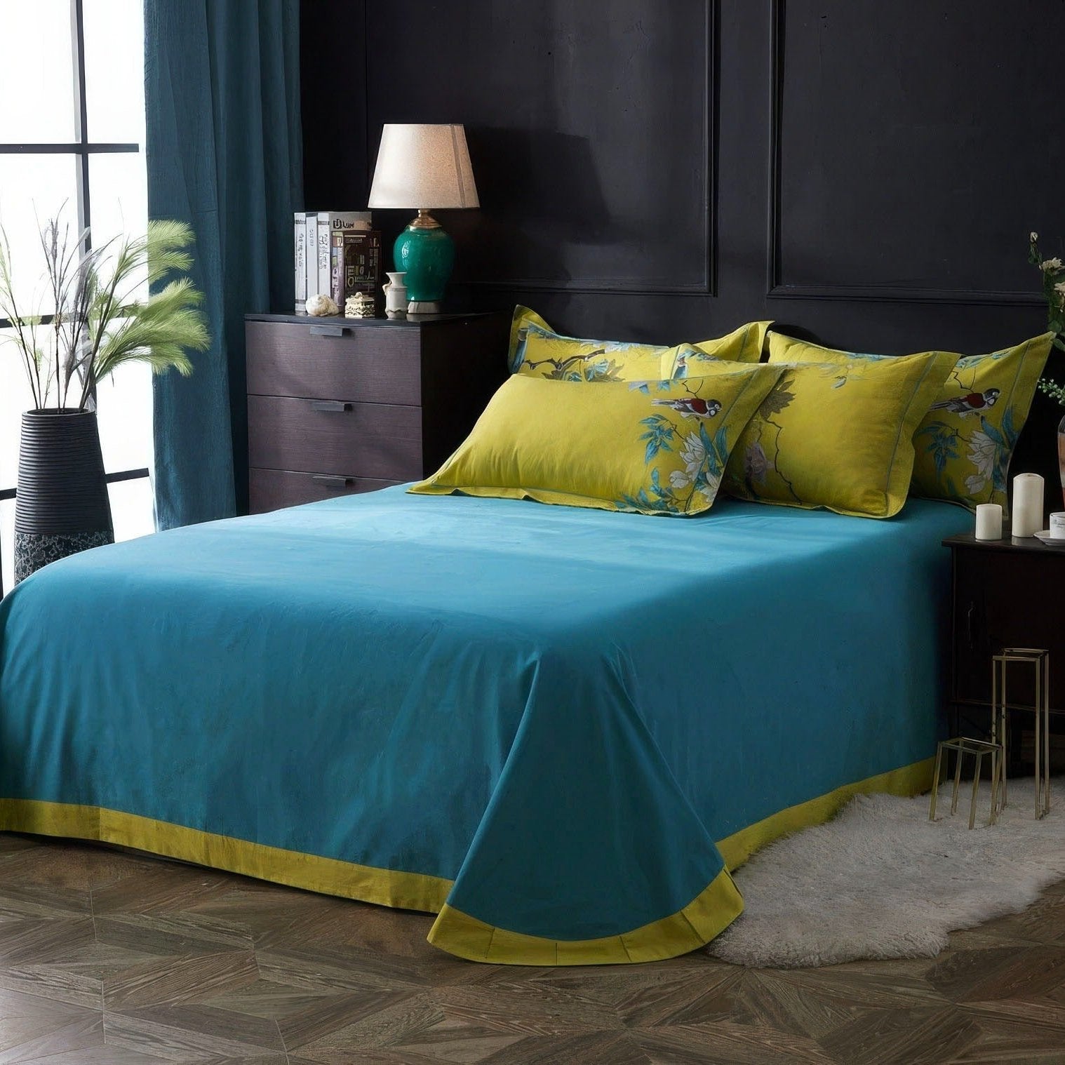 Vibrant bedroom with a teal bedspread accented by a yellow border, complemented by matching yellow pillowcases featuring embroidered birds and floral patterns. The dark wooden furniture, green vase lamp, and soft rug contrast with the teal curtains and a large window allowing natural light. A tall potted plant adds a fresh, natural element to the space.

