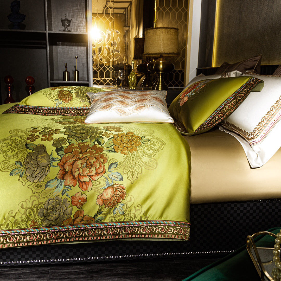 A close-up view of a luxurious lime green bedding set adorned with vibrant floral patterns in warm tones of orange, yellow, and green. The design is framed by intricate gold and red embroidered borders, adding an elegant touch. A white accent pillow with detailed golden embroidery complements the ensemble, while the room features a sophisticated decor with golden accessories, a table lamp, and a geometric reflective wall panel in the background.