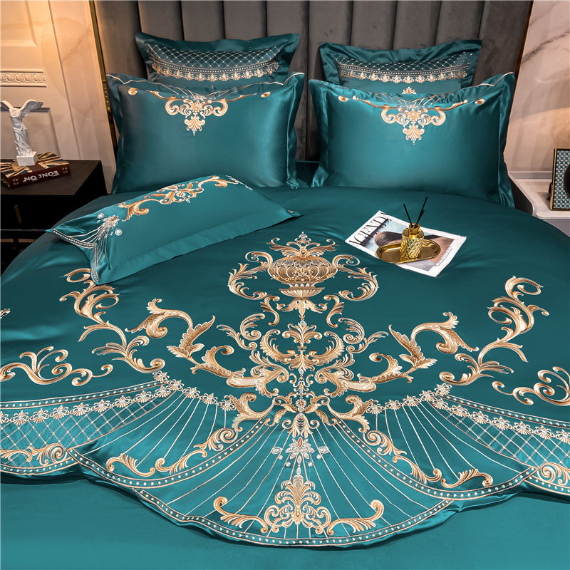 Green satin bedding set featuring gold baroque-style embroidery with ornate scrollwork and floral detailing. The intricate centerpiece design is symmetrically arranged, complemented by matching embroidered pillows and a decorative lumbar pillow. A tray holding a reed diffuser and magazine sits atop the smooth, lustrous surface, enhancing the luxurious presentation. The bed is set against a plush gray headboard with soft lighting and elegant bedside decor.

