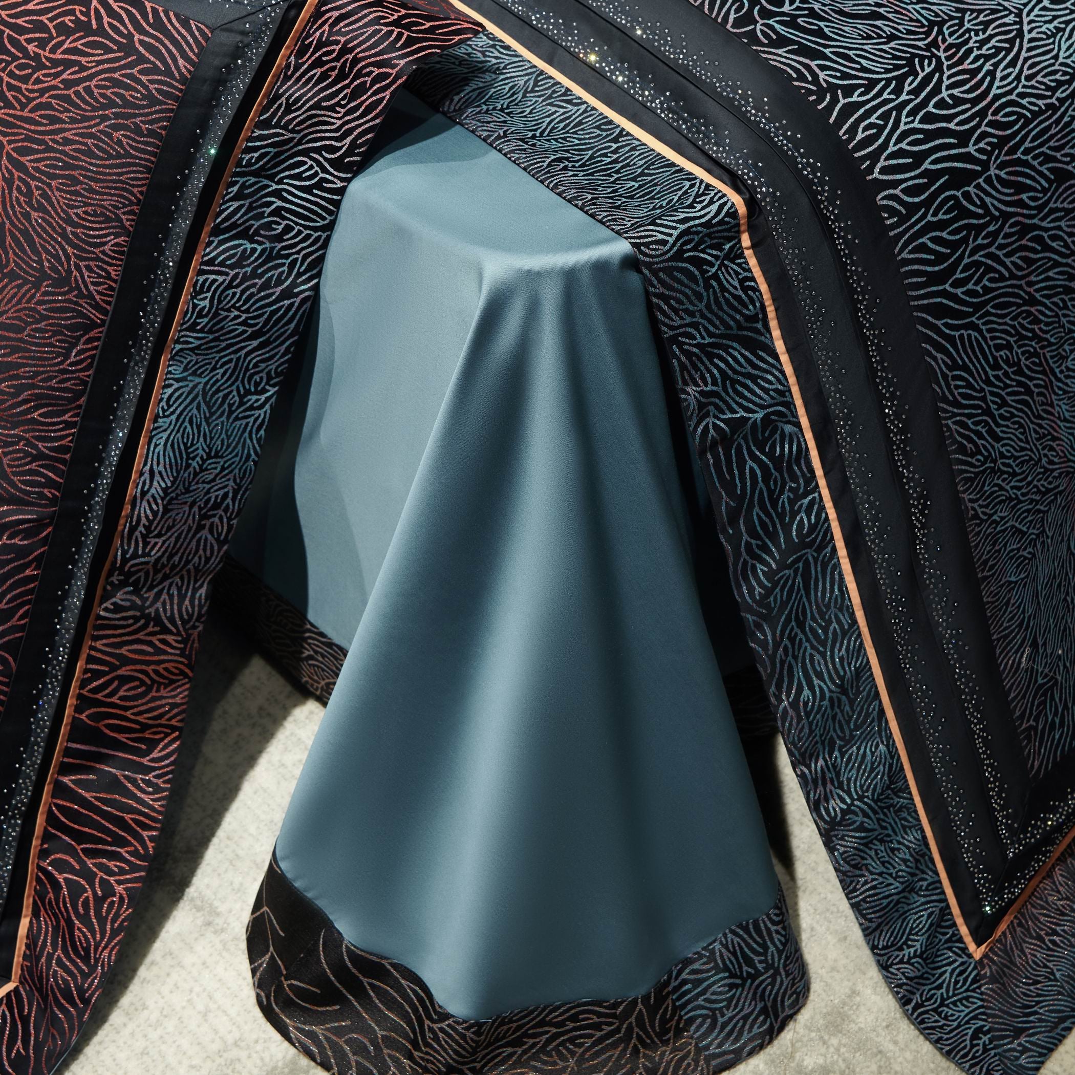 This close-up captures the luxurious drape of a high-end bedding set, showcasing a smooth, satin-like fabric in a refined teal hue. The bedspread features an intricate coral-inspired embroidery pattern flowing in rich red and blue tones on a deep black background, creating a striking visual contrast. The edges are delicately finished with shimmering crystal-like accents and a sleek copper-toned trim, adding a touch of elegance. 