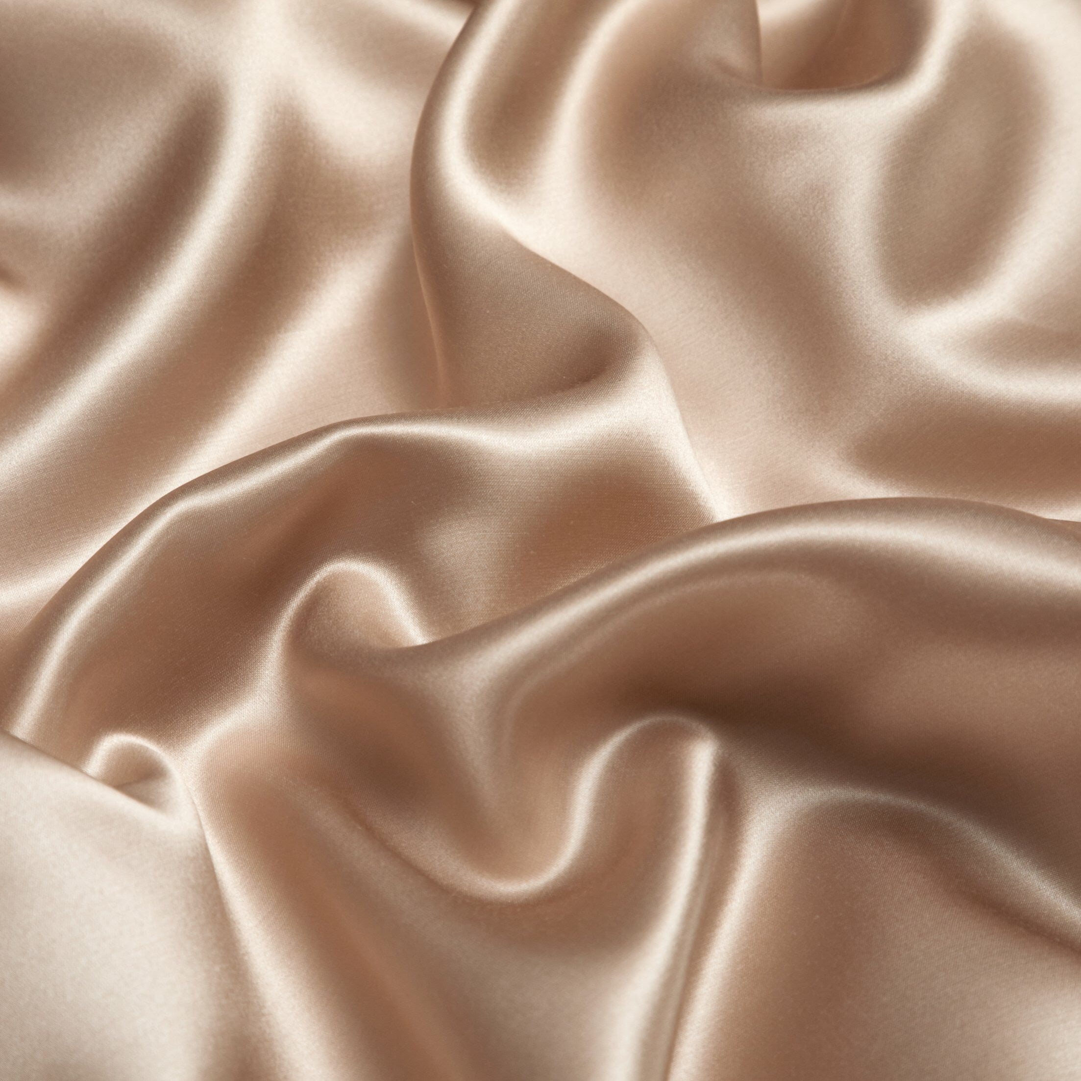 A detailed close-up of champagne gold silk fabric with a luminous, smooth texture. The material is luxuriously soft and showcases a gentle sheen, emphasizing its premium quality. The flowing, wavy drapes highlight the elegance and richness of the fabric.