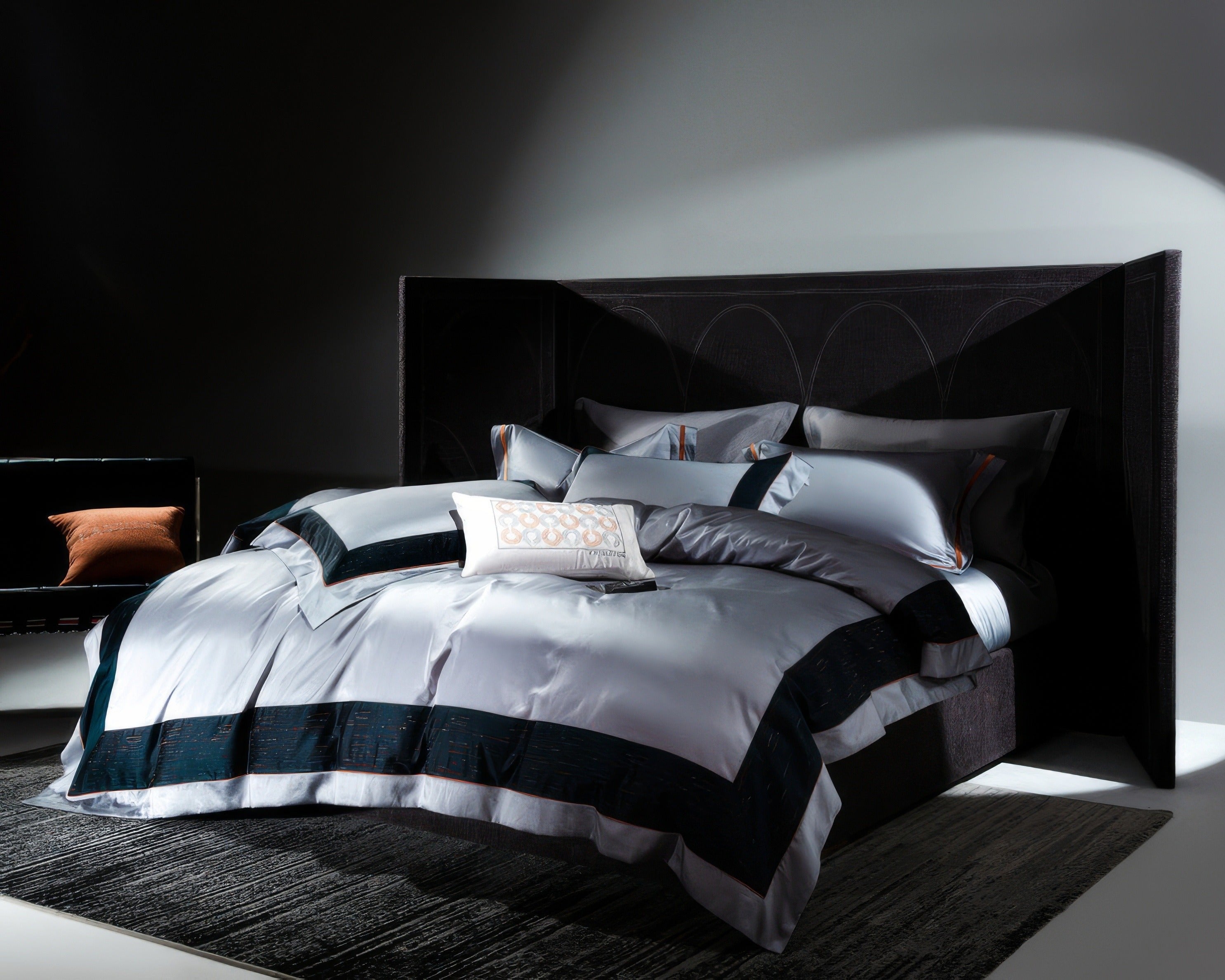 Modern bedroom featuring a luxurious bed with an oversized dark headboard and high-end bedding in silver and deep navy blue. The bedding includes a duvet cover with wide navy borders, matching pillow shams, and a decorative white cushion with subtle patterns. A dark carpet grounds the room, and a sleek black sofa with an orange pillow is placed to the side. Dramatic lighting casts soft shadows, highlighting the elegance of the bedding and creating a sophisticated, moody atmosphere.

