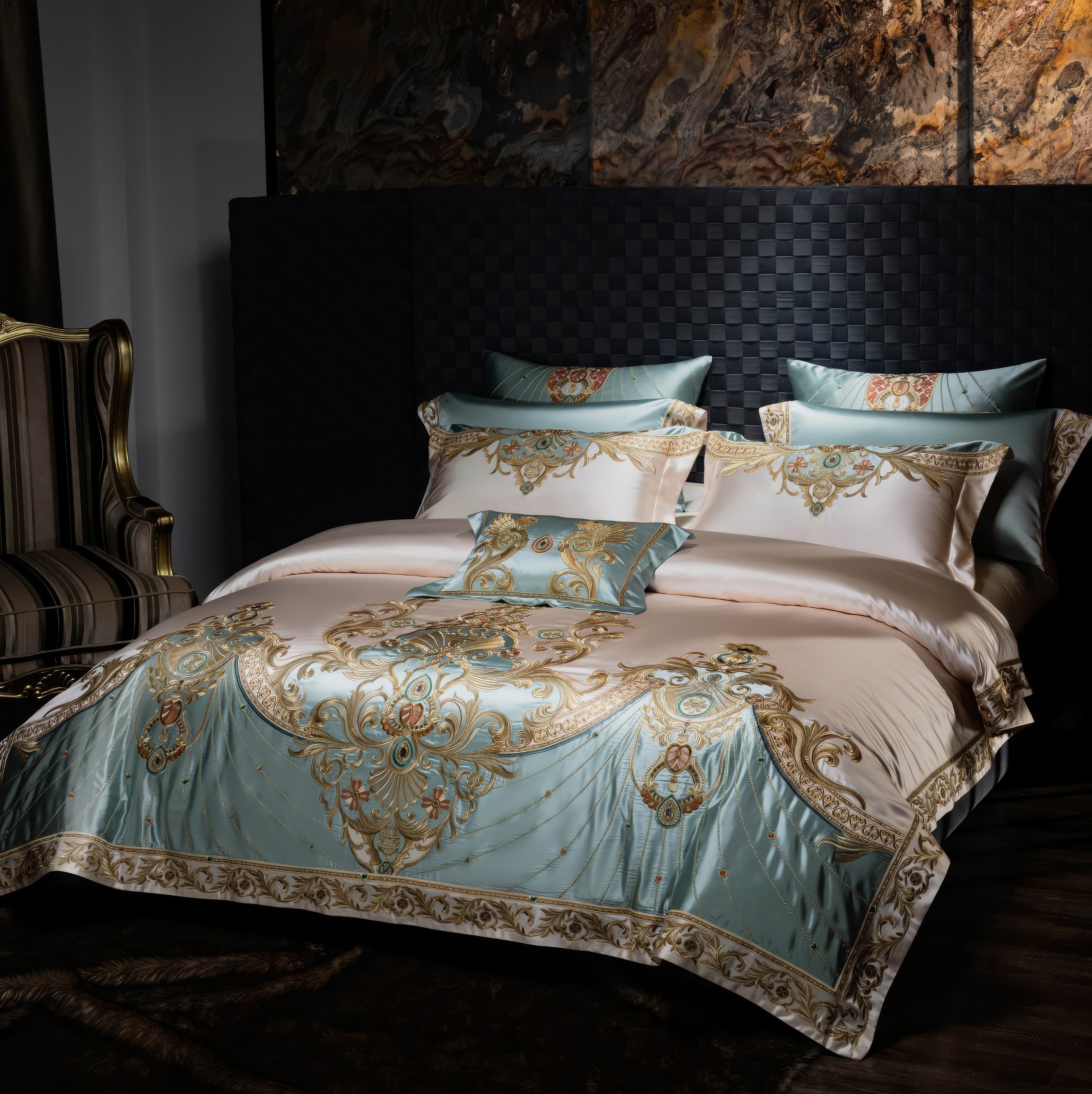 Elegant bedding set in light blue and cream tones with intricate gold baroque-style embroidery, paired with matching pillowcases and a decorative throw pillow, styled in a sophisticated bedroom with a striped armchair and a textured wall backdrop.