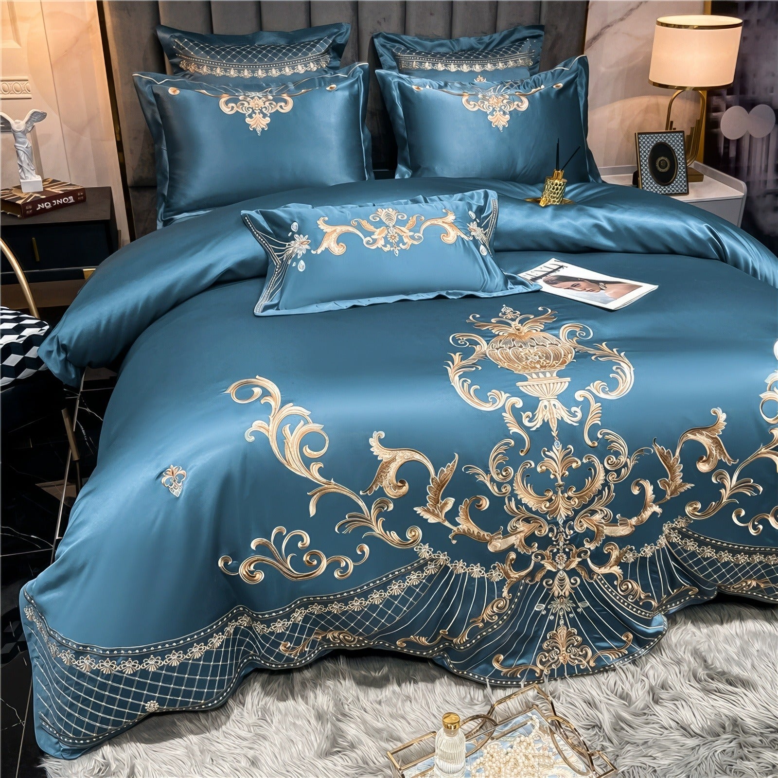 Elegant blue bedding set with intricate gold embroidery, including ornate scrollwork and floral patterns. Matching pillowcases and a decorative cushion complement the design, styled on a bed with a grey upholstered headboard. A glossy magazine and diffuser sit atop the bedding, while a plush grey rug adds texture to the floor.

