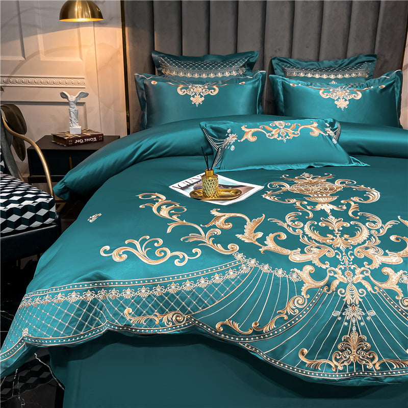 Green bedding set with intricate gold embroidery featuring ornate scrollwork, floral patterns, and a symmetrical baroque centerpiece design. The set includes matching decorative pillows and a lumbar pillow with coordinating embellishments. A reed diffuser and magazine rest on a tray atop the bed, while the scene is accented by soft lighting, a sculptural figure on a side table, and a geometric-patterned chair in the background.

