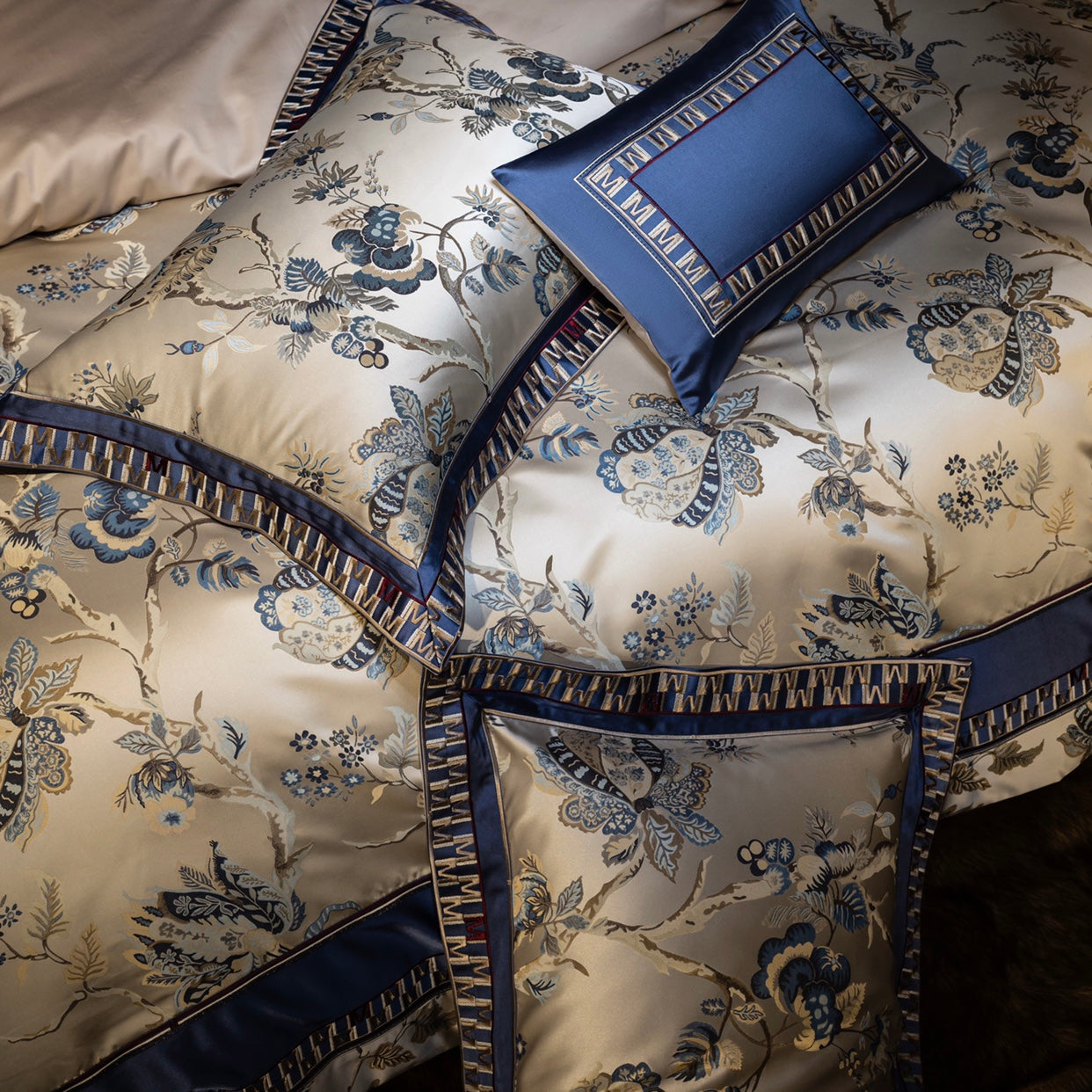 A detailed close-up of a luxury bedding set featuring a champagne base with intricate blue floral embroidery and ornate patterns. The design is framed by navy blue borders with geometric accents, adding depth and elegance. Coordinating pillowcases and a decorative cushion with a monogram-inspired "M" motif further enhance the cohesive and sophisticated aesthetic. The textured embroidery and satin finish emphasize the luxurious quality of the fabric, ideal for creating an opulent bedroom setting.