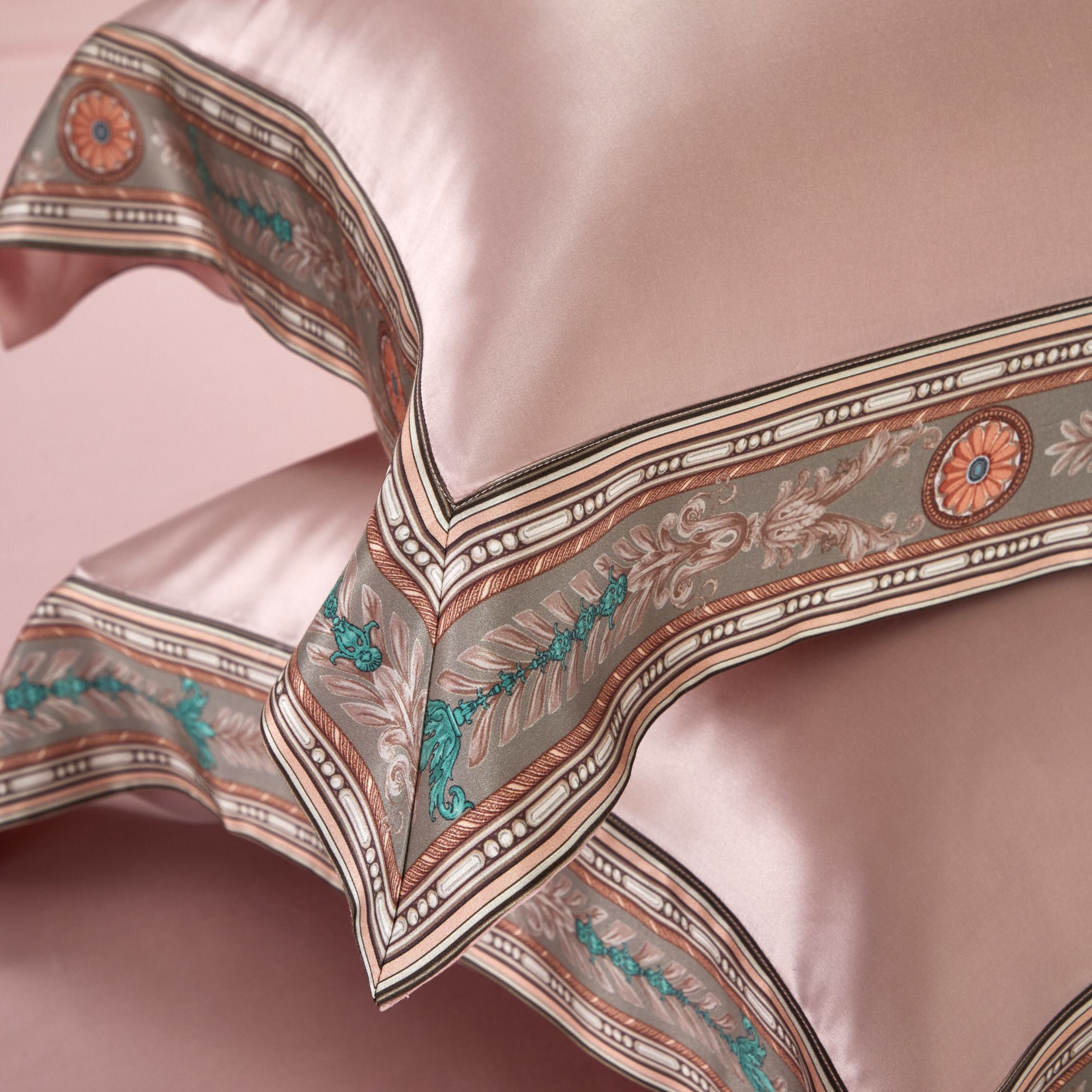 Close-up view of a blush pink pillowcase, highlighting its intricate ornamental border with floral and geometric motifs in soft earth tones and teal accents. The luxurious fabric showcases a smooth, glossy finish, enhancing the refined craftsmanship and opulent design details.