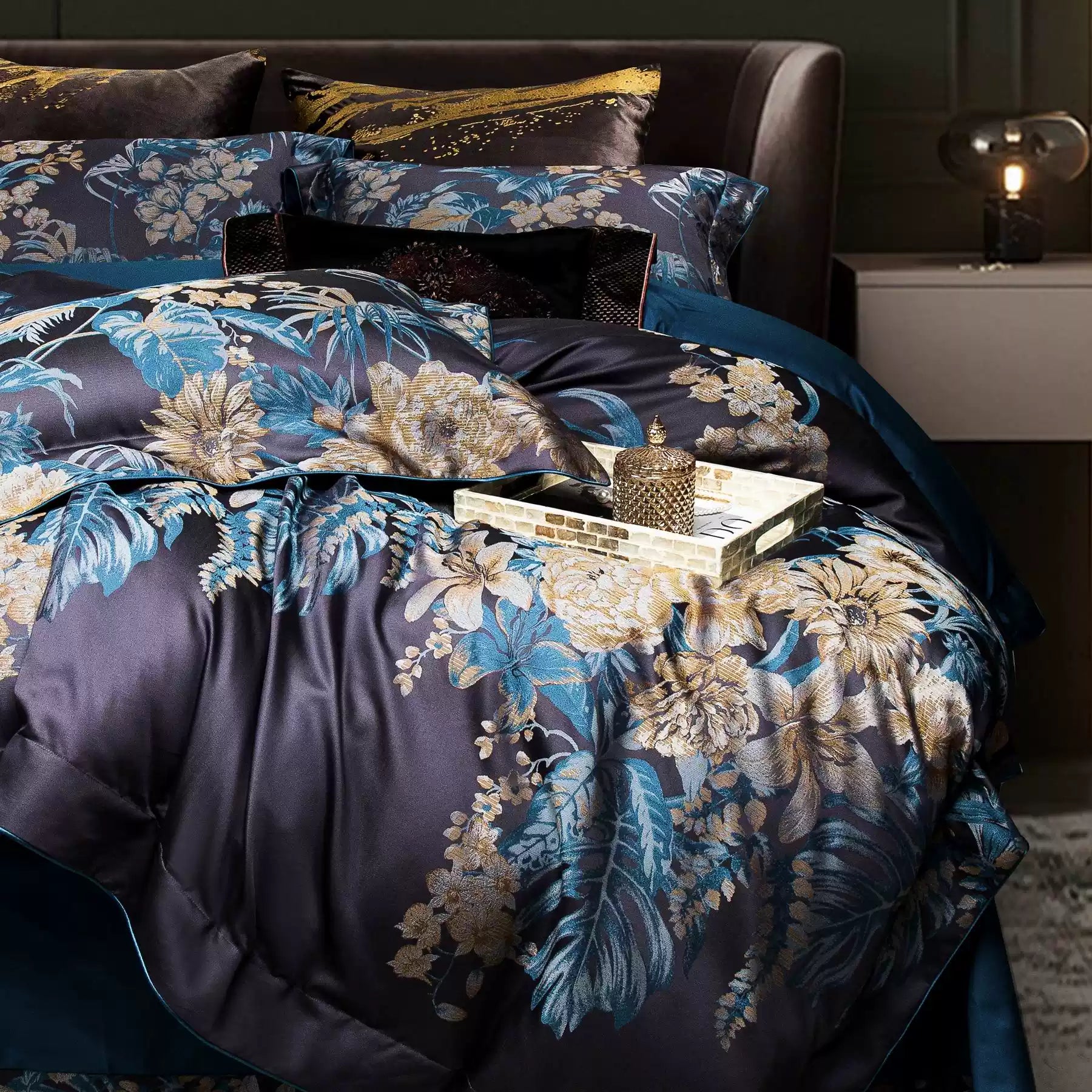 This image captures a close-up of the bedding set, showcasing its luxurious floral design against a navy backdrop. The vibrant gold and teal floral patterns are intricately woven, exuding elegance and sophistication. A sleek golden tray with a textured candle adds a touch of opulence to the setup, enhancing the overall aesthetic of the bedroom decor. The rich hues and metallic accents make this bedding set a standout piece, perfect for creating a statement in a modern or traditional bedroom setting.