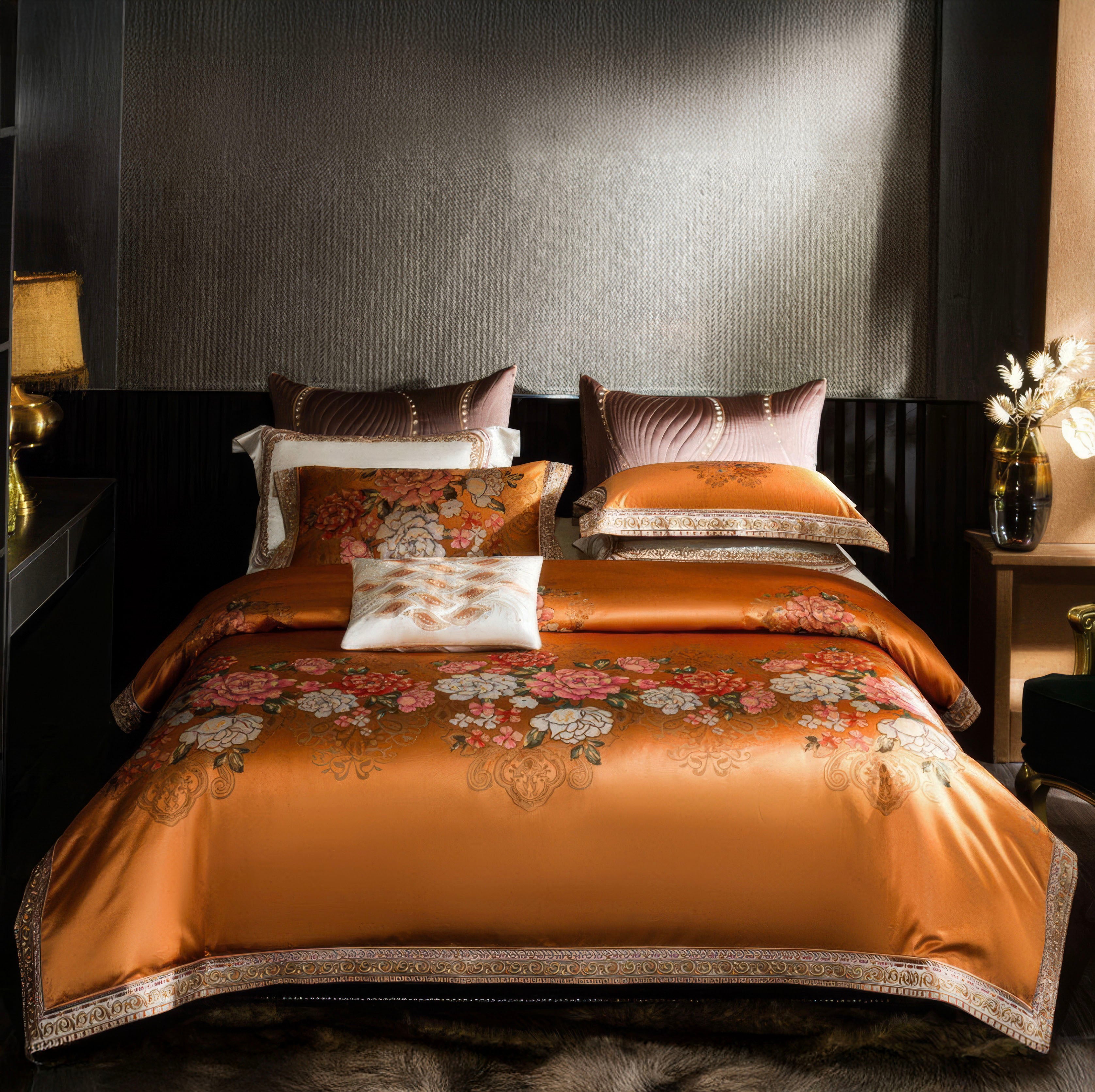 A luxurious bedroom setup featuring a vibrant orange bedding set adorned with intricate floral patterns, exuding elegance and charm. The duvet cover, complemented by matching pillowcases and accent pillows, boasts ornate golden and cream detailing, enhancing its regal appeal. The backdrop includes a sophisticated textured wall, ambient lighting from a gold-toned table lamp, and a decorative vase with dried flowers on a wooden console table. The warm and cozy atmosphere is completed by a fur-style area rug, 