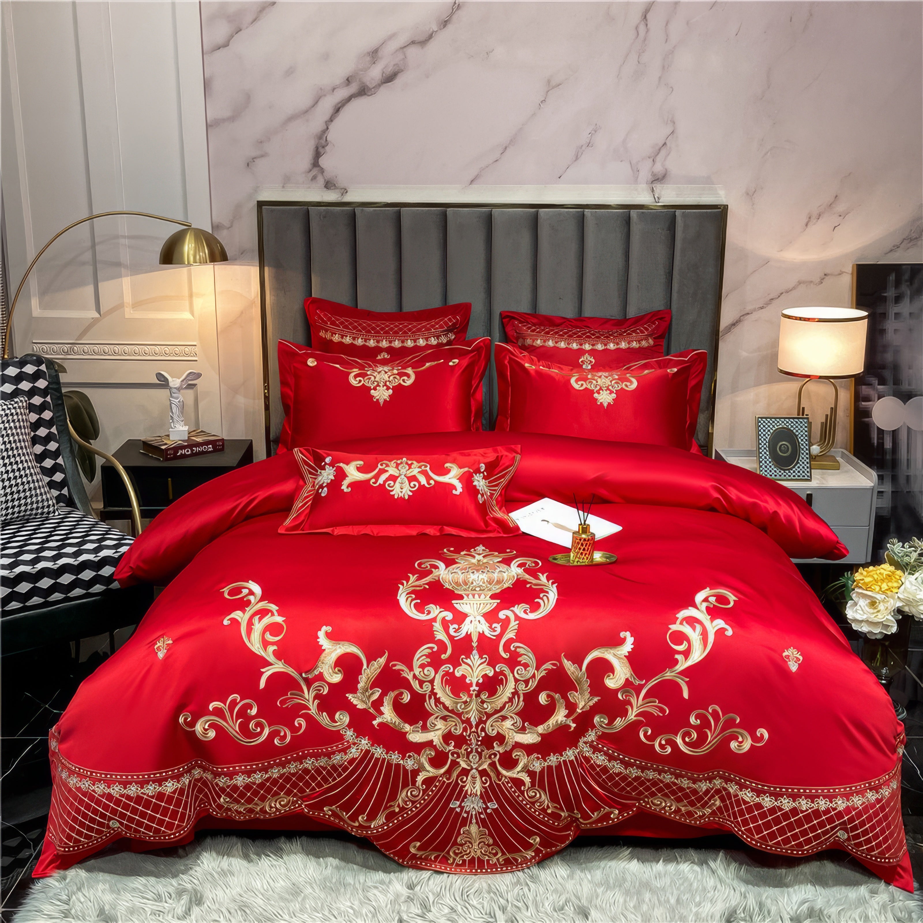 Red Egyptian cotton bedding set adorned with intricate gold embroidery featuring ornate scrollwork and floral motifs. The set includes matching pillowcases and a duvet cover, all showcasing the luxurious design. The vibrant red hue and soft sheen of the fabric highlight the premium quality of Egyptian cotton, arranged elegantly against a grey upholstered headboard with a marble wall backdrop and a soft white rug beneath.

