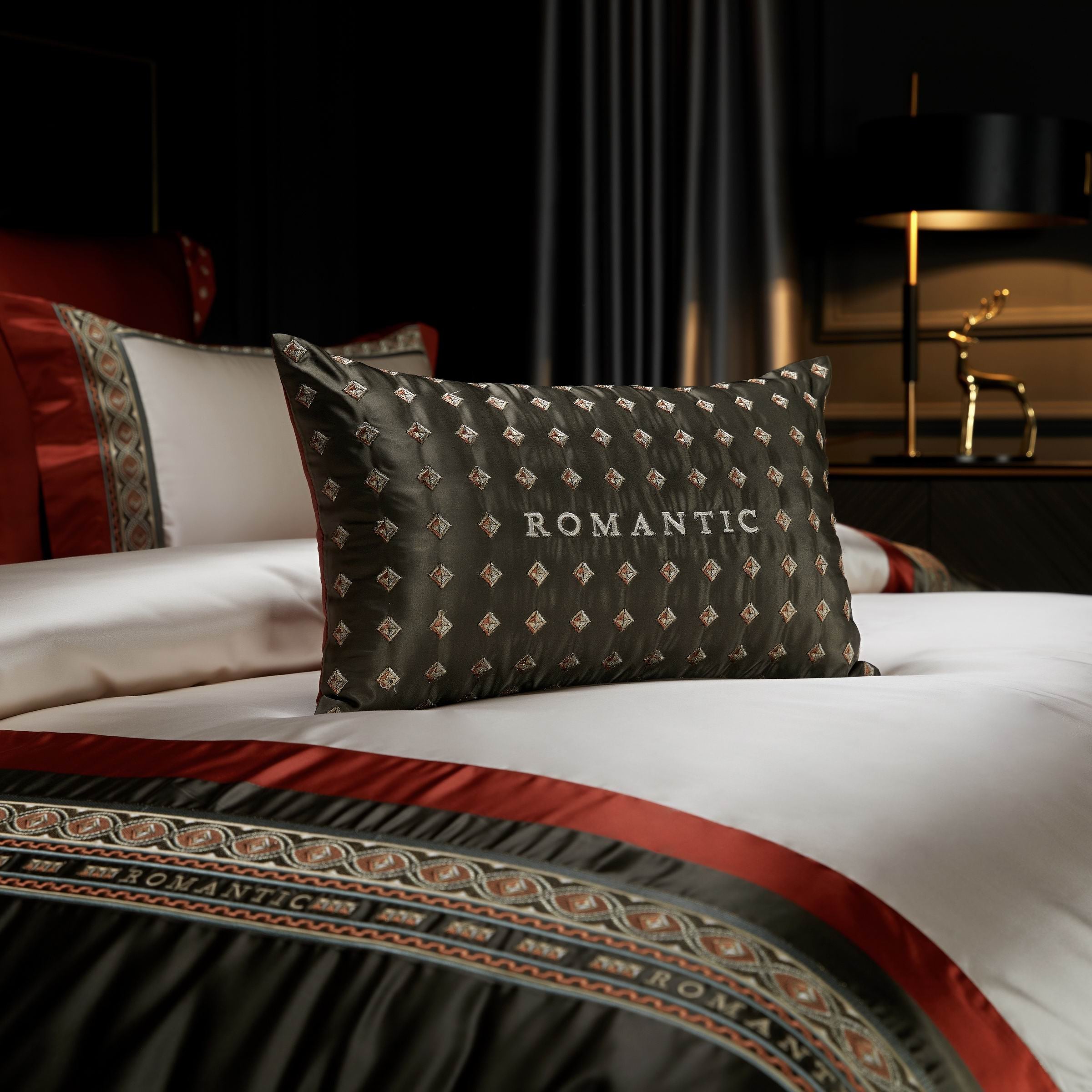 A close-up view of a decorative bolster pillow placed on a luxurious bedding set. The pillow, crafted in deep olive green fabric, features a subtle diamond-shaped pattern with delicate golden accents and the word "ROMANTIC" elegantly embroidered in bold white lettering. The surrounding bedding showcases intricate border designs in olive green, crimson red, and ivory tones, complementing the sophisticated theme. In the background, soft lighting illuminates a modern bedside table adorned with a golden deer fi