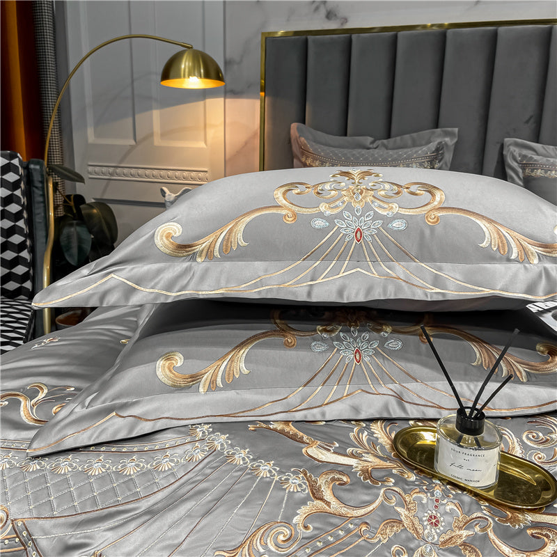 Grey bedding set with detailed gold and silver embroidery, featuring symmetrical scrollwork and floral motifs on pillowcases and duvet cover. The fabric has a smooth, satin-like sheen, complemented by elegant piping along the edges. A gold tray with a black reed diffuser sits atop the bedding for a luxurious touch.

