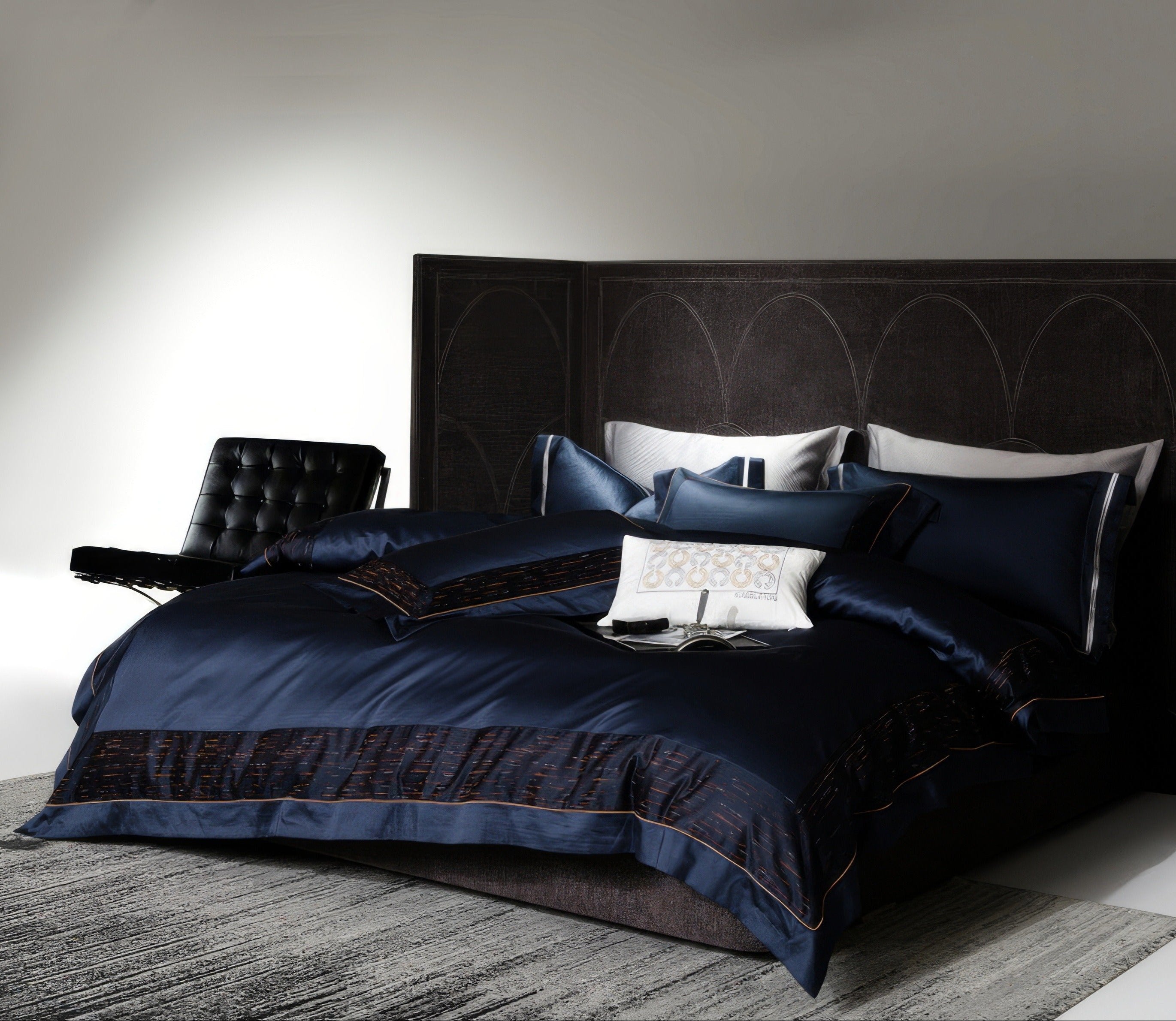 Modern bedroom featuring a dark navy blue luxury bedding set with subtle textured black and red borders. The set includes multiple pillow arrangements, a white decorative cushion at the center, and a plush duvet. A black leather chair sits beside the bed, complementing the dark arched-panel headboard, creating a sleek and sophisticated ambiance.

