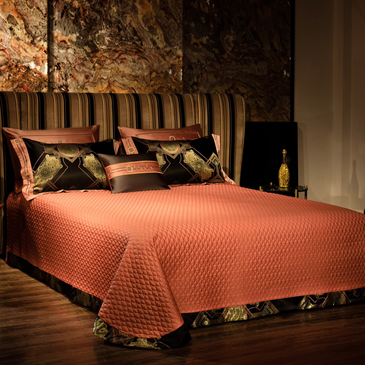 A luxurious bedroom scene with a copper-colored quilted bedspread covering the bed, paired with black pillows featuring intricate golden geometric designs and copper trim. The striped headboard and marble-textured wall in the background enhance the rich and elegant ambiance, while a gold-detailed decorative bottle on the side table completes the sophisticated setting.