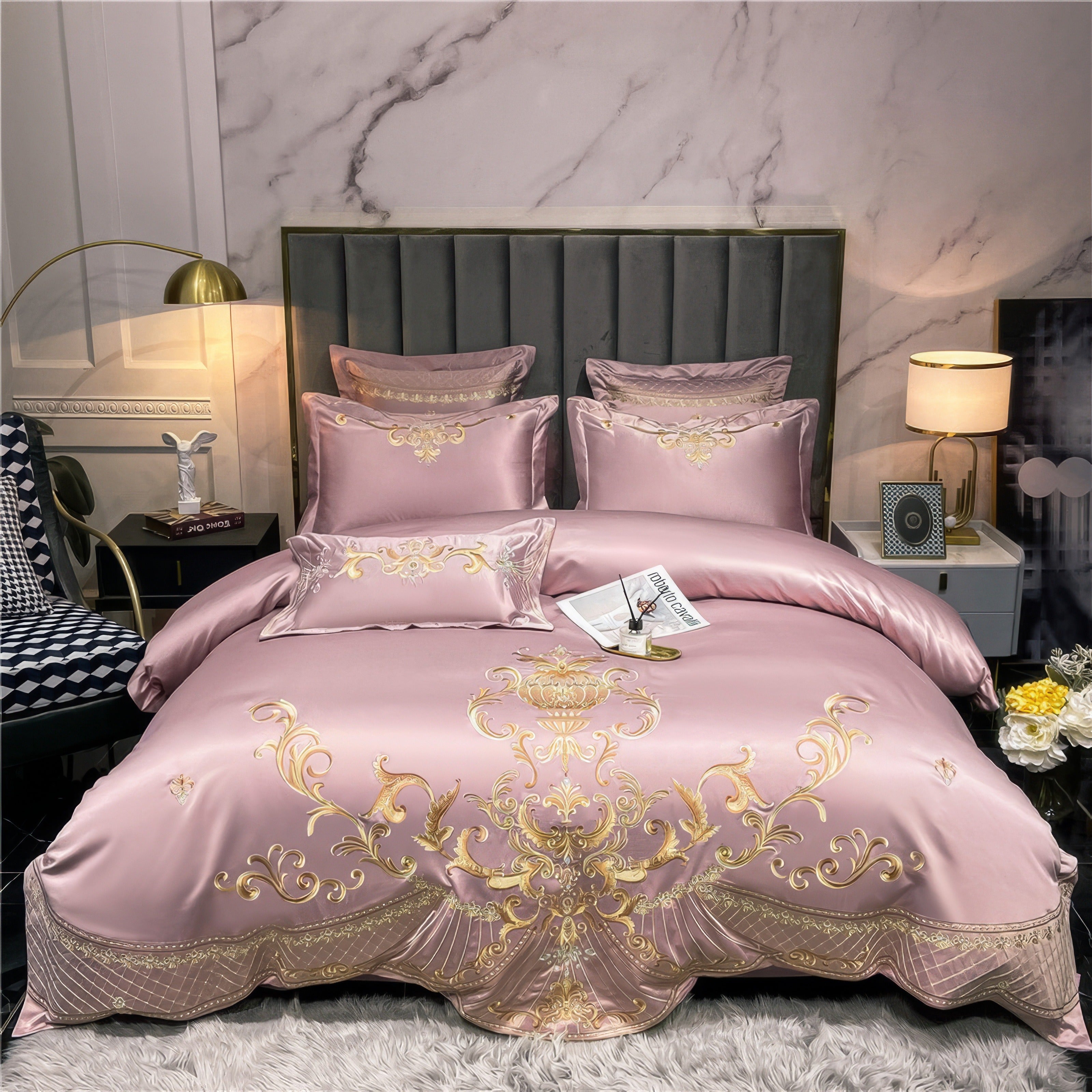 Luxurious pink satin bedding set with gold embroidery in a regal pattern on the duvet cover. Matching pillowcases and decorative cushions are arranged against a plush gray headboard. The scene is lit by soft ambient lighting from a gold floor lamp and a table lamp, with a marble accent wall in the background.

