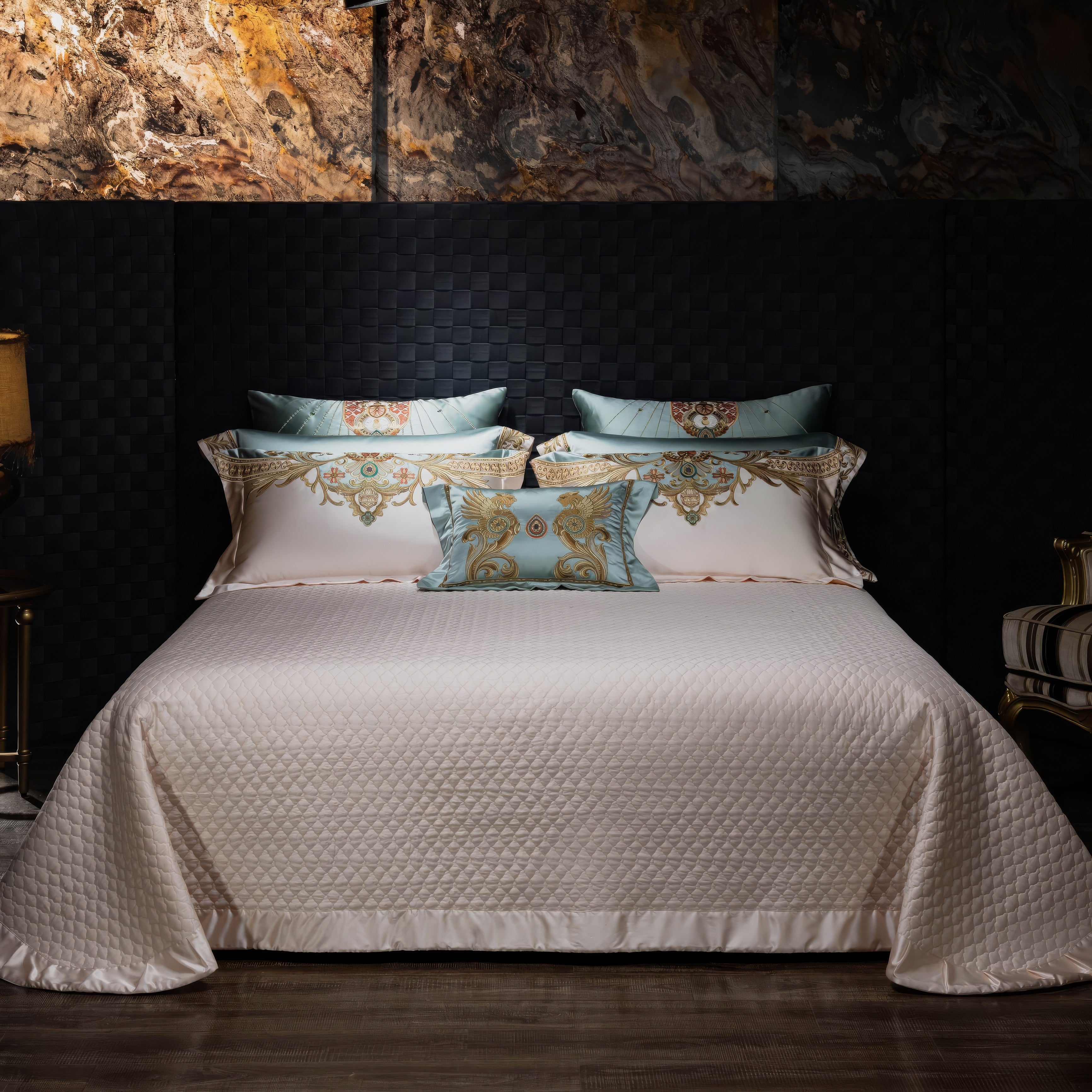 Elegant bedding set featuring a cream quilted bedspread and matching pillowcases with light blue and gold baroque-style embroidery, styled in a luxurious bedroom with a textured black wall and marble accents