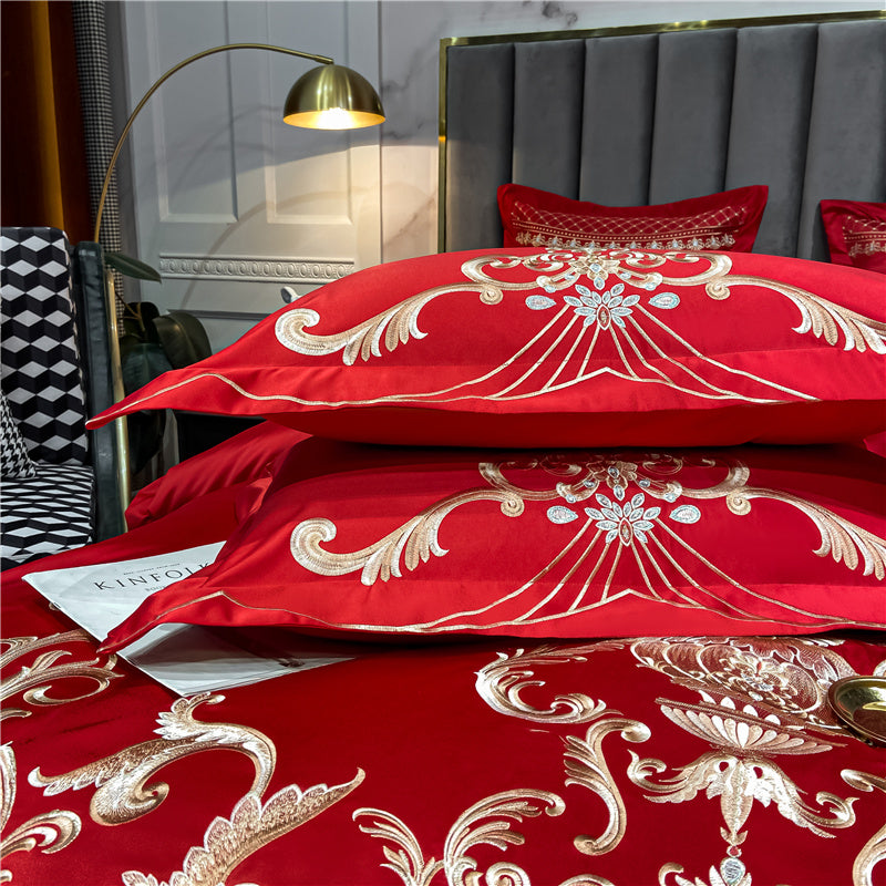 Close-up of a red Egyptian cotton bedding set with detailed gold embroidery on pillowcases and the duvet. The design features ornate scrollwork and floral motifs, with shimmering threads creating an elegant, luxurious look. The pillows are stacked neatly, showcasing the bold red color and symmetrical embroidery. The bedding rests on a glossy red surface, accompanied by a glimpse of a magazine and a gold tray for added sophistication.

