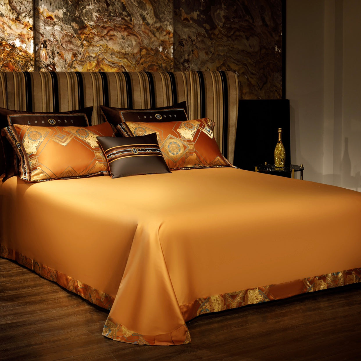 An orange bedspread with a smooth, sleek finish draped elegantly over a bed, paired with matching pillowcases featuring intricate gold and blue geometric patterns and dark brown accents. The setup is complemented by a striped upholstered headboard and gold-tone accessories, creating a rich and luxurious bedroom design with a warm, opulent ambiance.