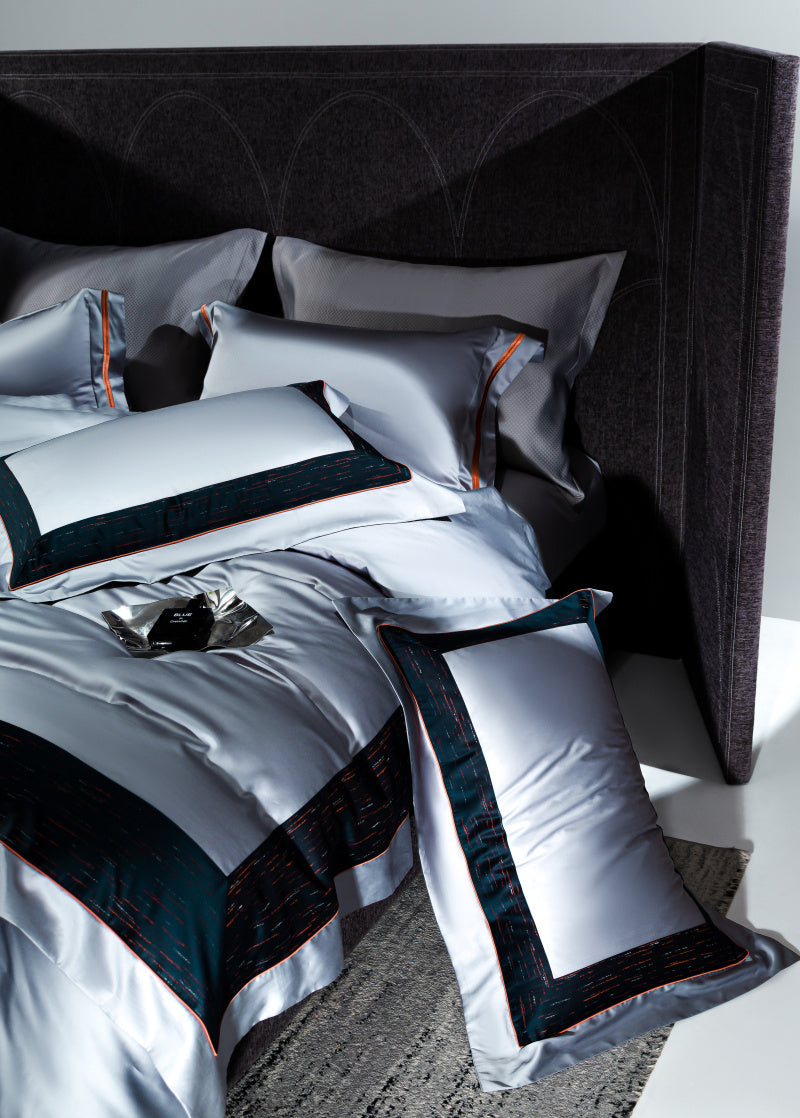 Luxurious bedding set in a contemporary design featuring silver fabric with bold black and red borders on pillowcases and a duvet cover. The arrangement includes neatly layered pillows against a dark, textured headboard, with a decorative black and silver accessory on the bed for added sophistication.

