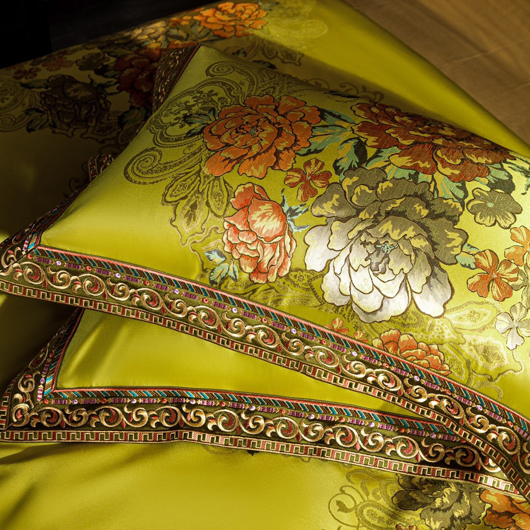 A close-up view of lime green pillowcases showcasing a vibrant floral print in shades of orange, red, and cream, framed with ornate gold embroidery and colorful geometric accents. The intricate border design and rich detailing enhance the opulent appearance of the fabric, while the silky texture adds a luxurious touch to the bedding ensemble.