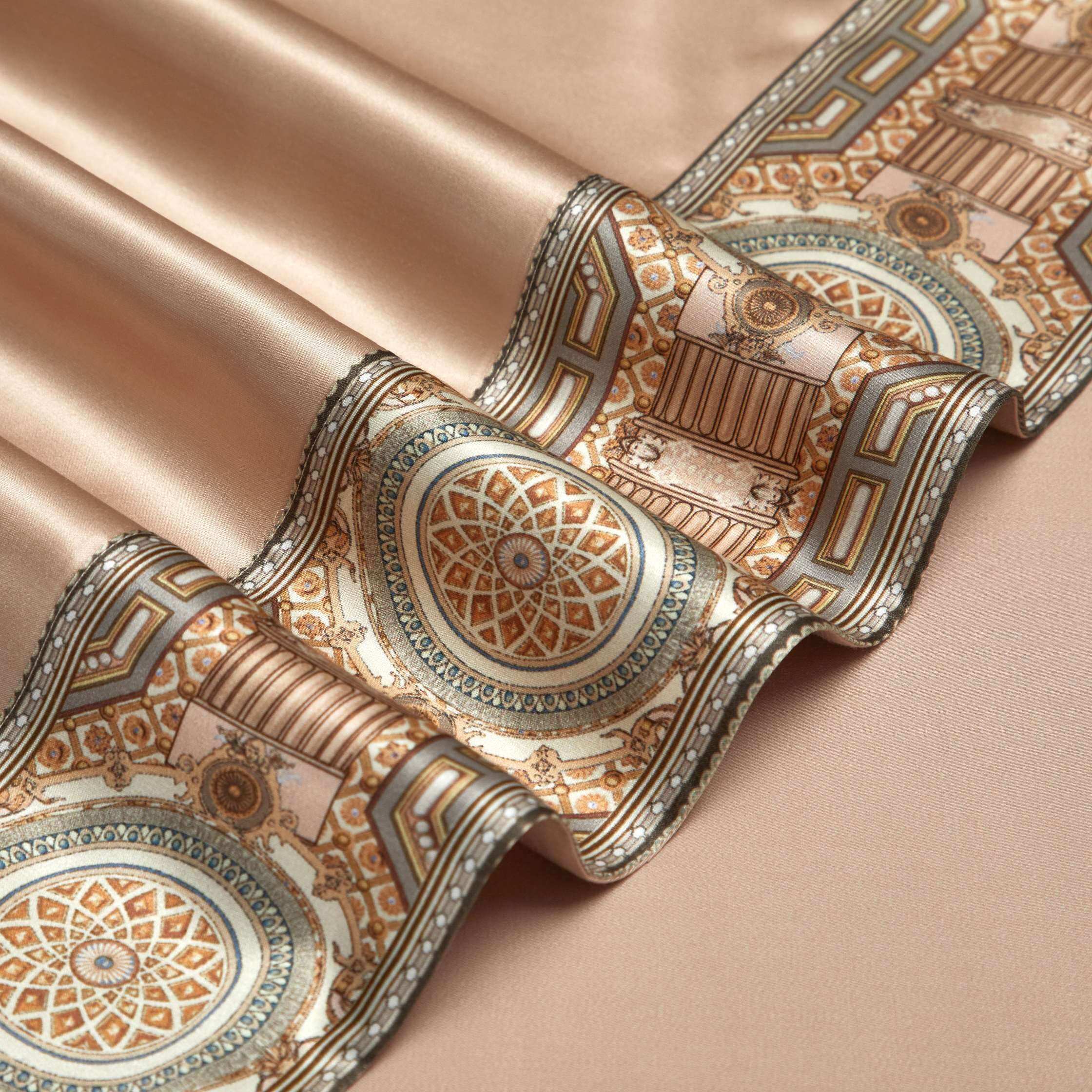 A close-up of a champagne gold silk fabric adorned with an intricate border design. The border features an elegant geometric pattern with circular medallions in gold, bronze, and teal, surrounded by ornate architectural details, adding a sophisticated and luxurious touch. The smooth texture of the silk is highlighted by its soft sheen and precise detailing.