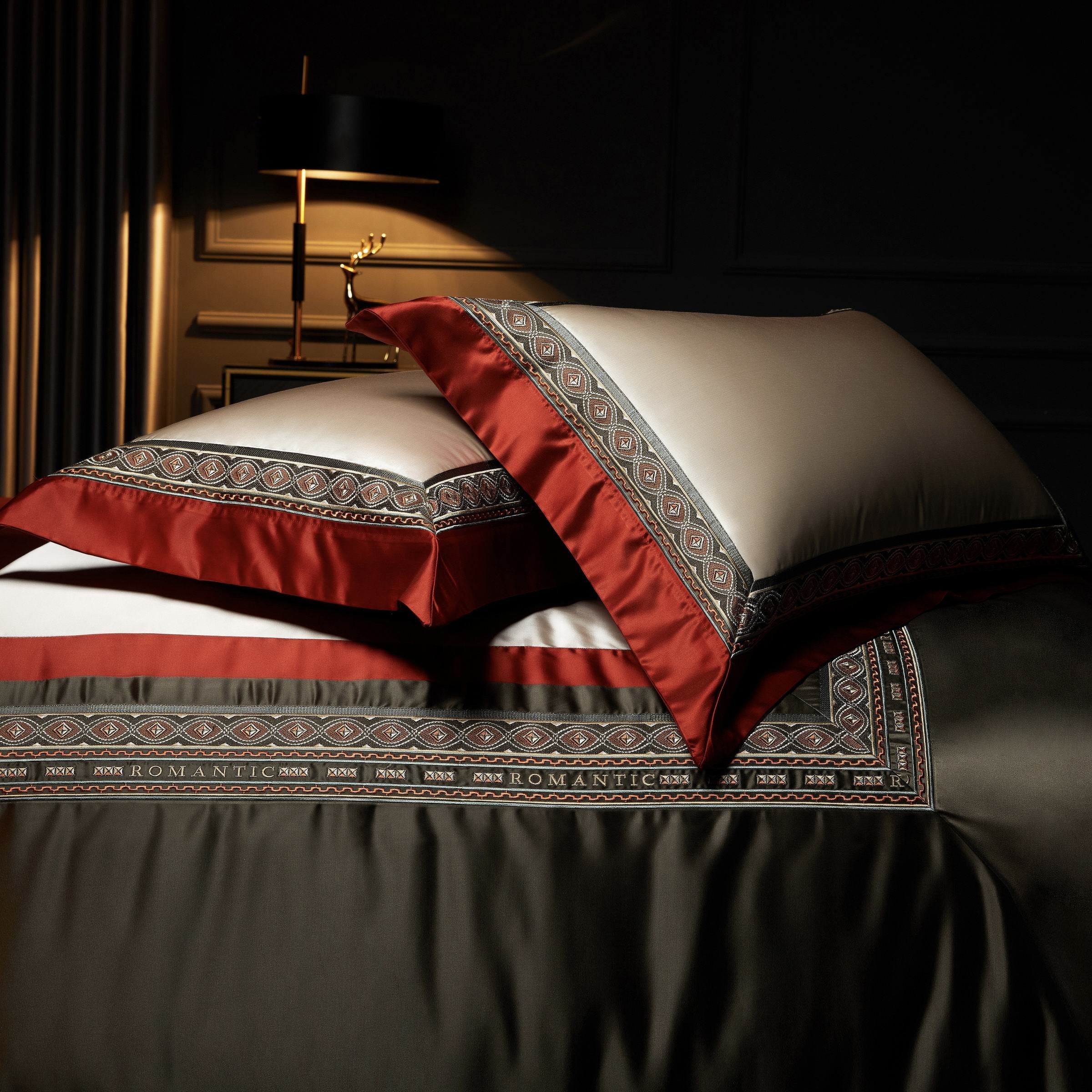 A close-up view of an elegant bedding ensemble showcasing ivory pillowcases framed with bold crimson red edges, accentuated by intricate olive green and gold embroidered patterns. The luxurious design extends to the bedspread, which features a matching border with the word "ROMANTIC" subtly integrated into the decorative details. A softly lit background with a modern black lamp and gold-toned decor adds to the opulent ambiance of the scene.