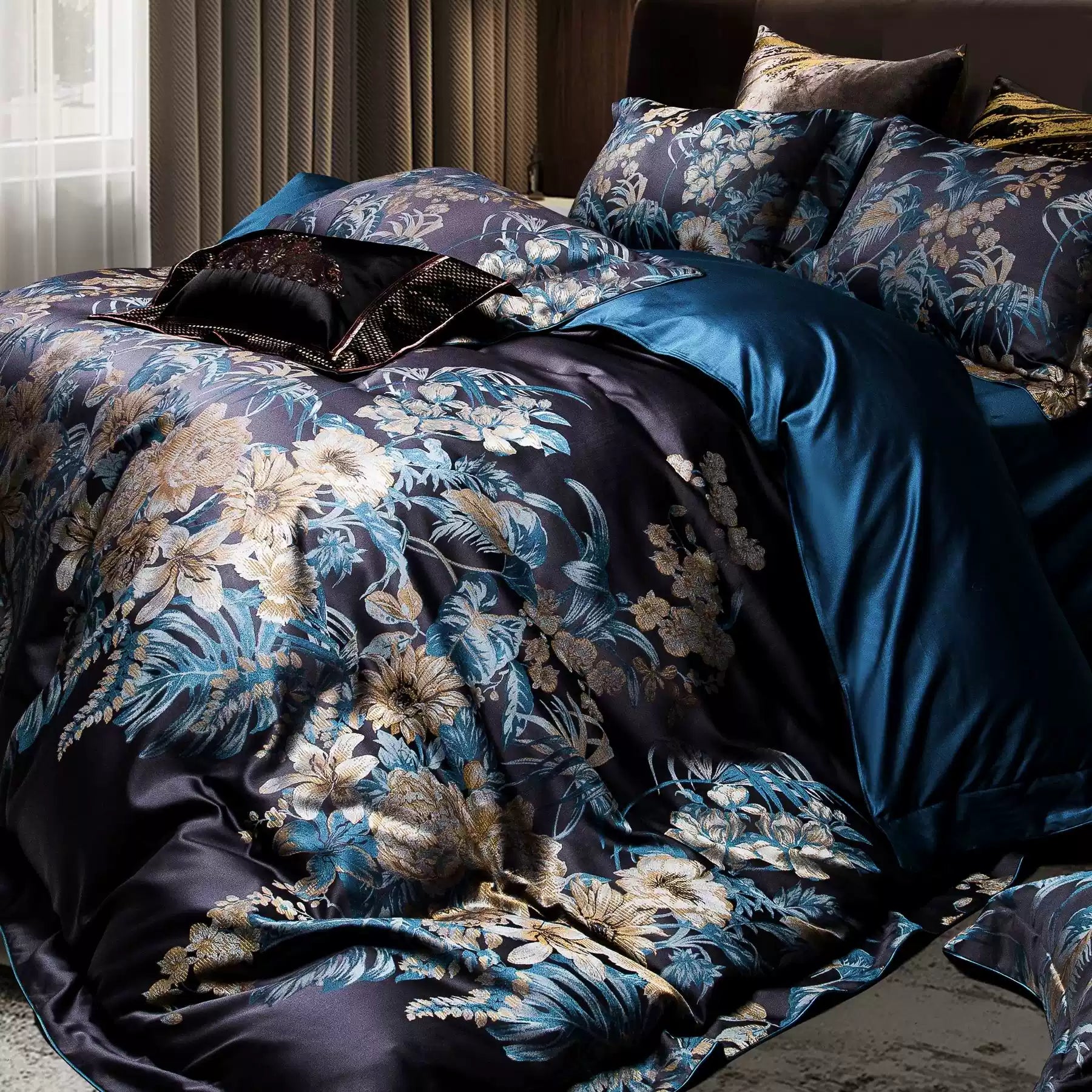 A close-up of a luxurious bedding set featuring a captivating floral design in vivid blue and gold hues on a dark background. The intricate patterns exude sophistication, complemented by a rich teal bedsheet and a black accent pillow with metallic details. The satin-like fabric enhances the bedding's opulence, reflecting light beautifully. The image showcases the fine craftsmanship and premium material, perfect for a refined bedroom aesthetic.