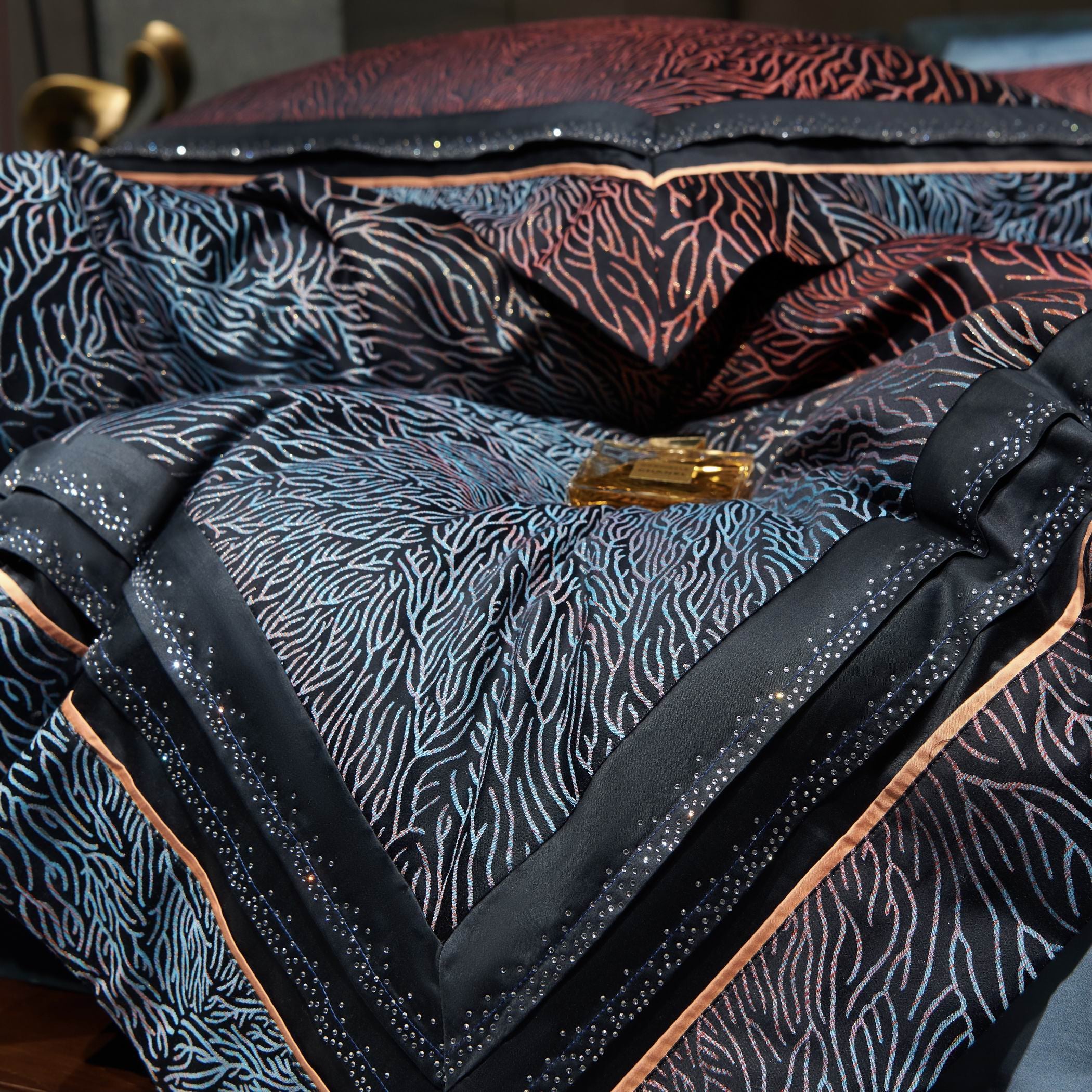 This close-up highlights the luxurious details of a high-end bedding set, showcasing a mesmerizing coral-inspired pattern embroidered in shades of blue and red against a sleek black background. The intricate embroidery seamlessly flows across the fabric, enhanced by shimmering crystal-like accents along the border that add a touch of opulence. A polished copper-toned trim outlines the design, elevating its contemporary elegance. 