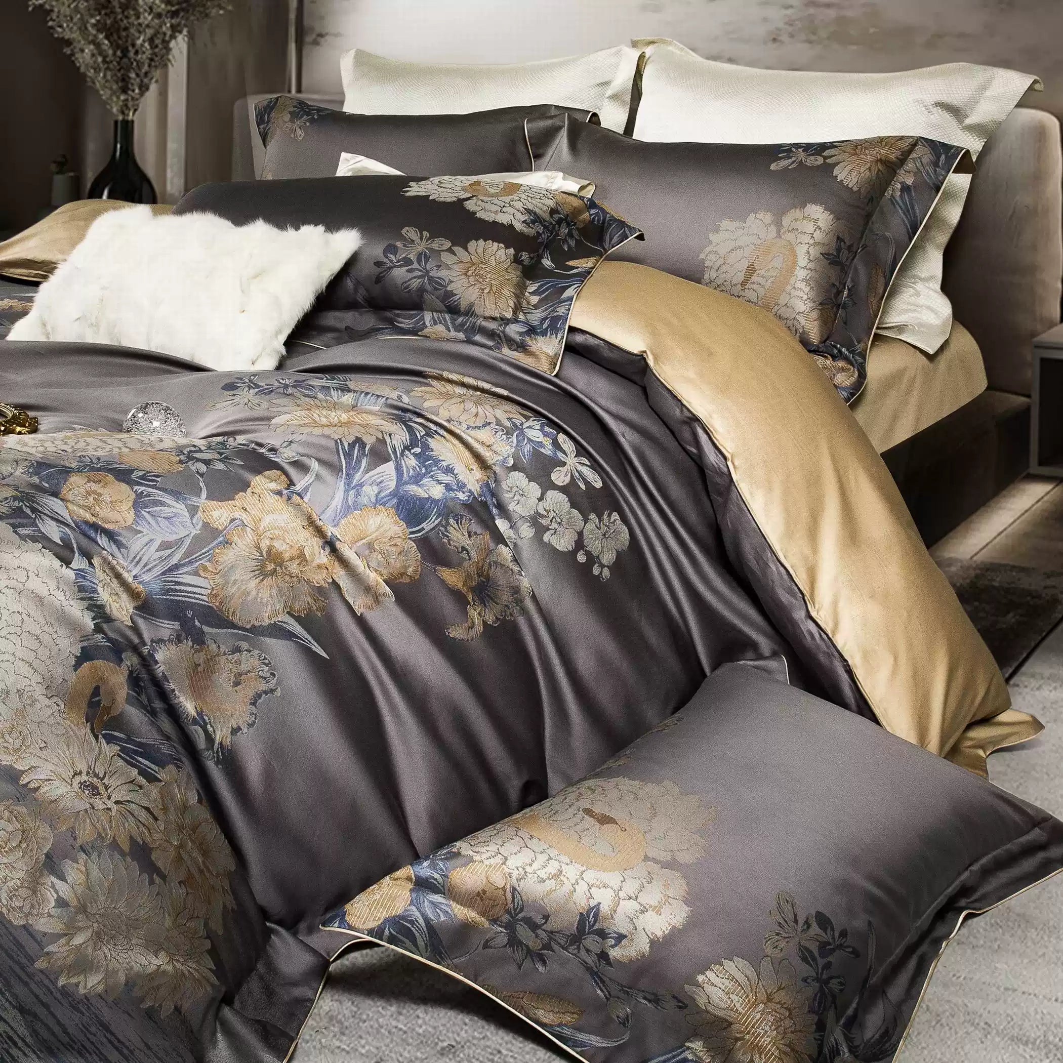 A close-up of a luxurious bedding set showcasing a dark gray duvet with elegant floral patterns in gold and blue. The bedding is complemented by matching pillowcases and a gold-tone bed sheet that adds warmth and contrast. A fluffy white decorative pillow sits atop the bed, enhancing its sophisticated and inviting appeal.