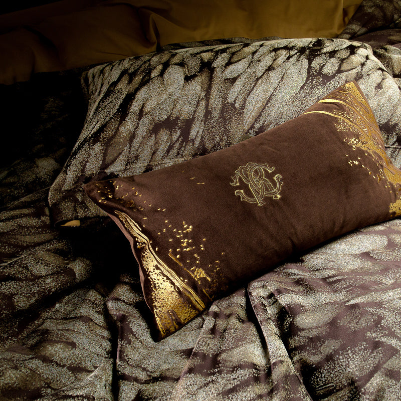 A close-up view of a luxury bedding set featuring a rectangular velvet throw pillow in deep brown with gold accents. The pillow is adorned with an embroidered monogram "R" surrounded by ornate detailing and abstract golden splashes, exuding elegance. The background showcases a complementary duvet cover with a textured pattern in shades of brown and gold, creating a harmonious and opulent look. The rich tones and intricate designs enhance the luxurious appeal of the bedding ensemble.