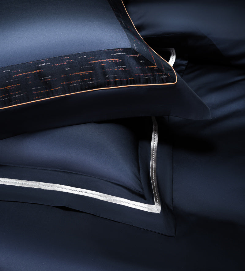 Close-up of navy blue bedding with intricate fabric detailing, featuring orange streak patterns and elegant silver piping along the pillow edge. The luxurious texture and sharp contrast of colors highlight the premium quality of the material.

