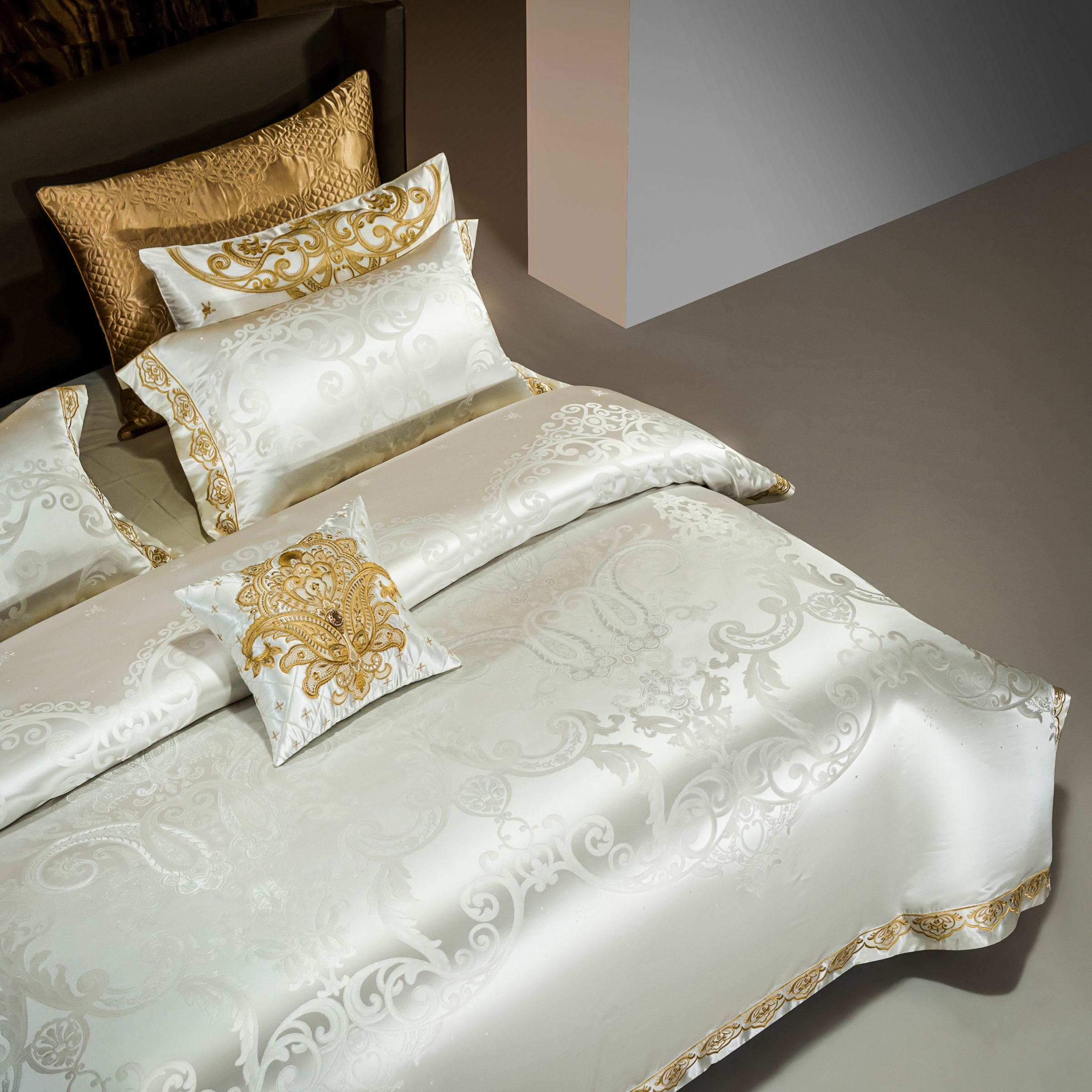 An overhead view of a luxurious bedding ensemble in white, featuring intricate gold jacquard patterns and embroidery. The duvet cover is complemented by matching pillowcases and a decorative throw pillow, all displaying opulent detailing. Golden accent pillows add richness to the overall design, perfect for an elegant and sophisticated bedroom decor.

