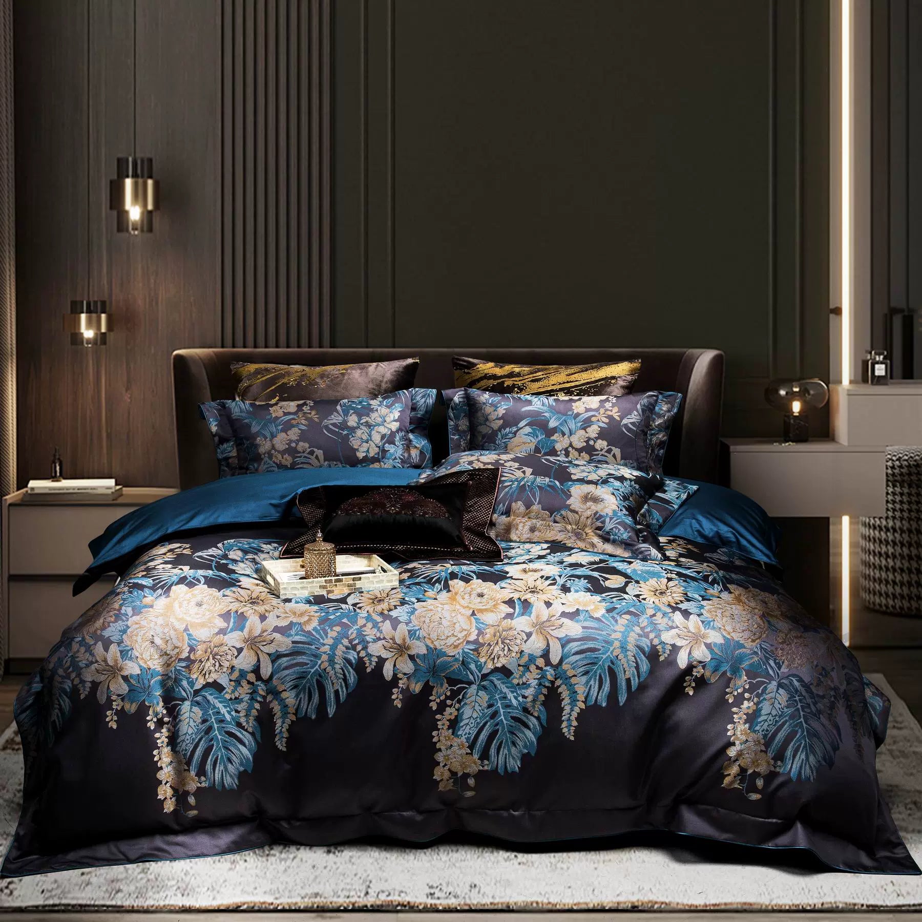 A luxurious bedding set in a dramatic dark palette featuring bold floral patterns in shades of blue and gold. The bed is adorned with multiple pillows showcasing intricate designs, complemented by a rich teal sheet that adds depth to the ensemble. The black accent pillow with metallic embellishments brings a touch of sophistication. The room's contemporary ambiance is enhanced by warm wood paneling, minimalistic pendant lighting, and modern bedside furniture, creating a cozy yet elegant retreat.