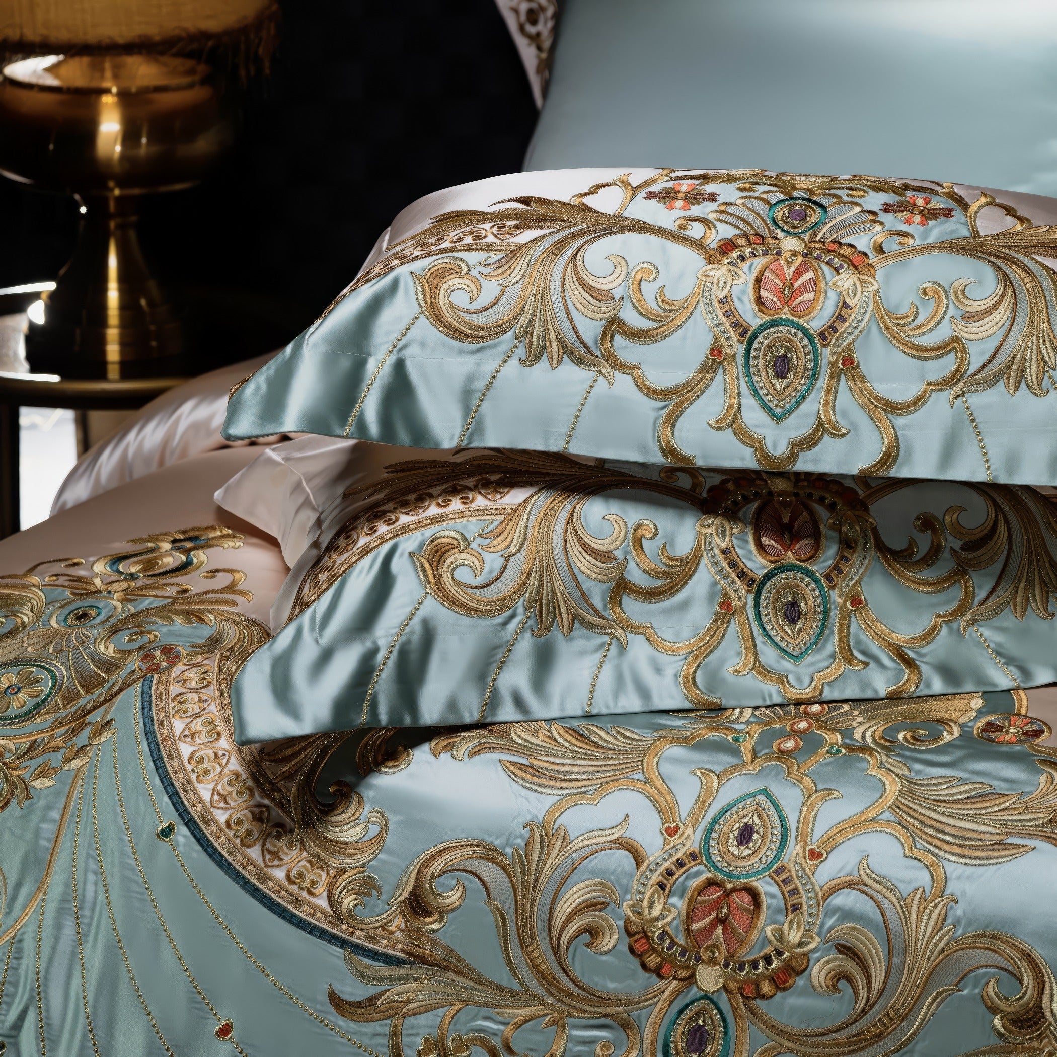 Stacked pillowcases in light blue satin with intricate gold embroidery featuring baroque-inspired patterns and jewel-tone accents, styled on a luxurious bedding set with a golden lamp in the background.