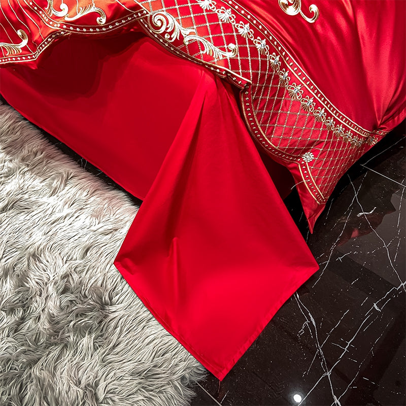 Red Egyptian cotton bedding with a solid red flat sheet cascading onto a soft, plush grey rug. The edge of the duvet showcases intricate gold embroidery in lattice and floral patterns, contrasting beautifully against the vibrant red fabric. The polished black marble floor reflects the luxurious details of the bedding.

