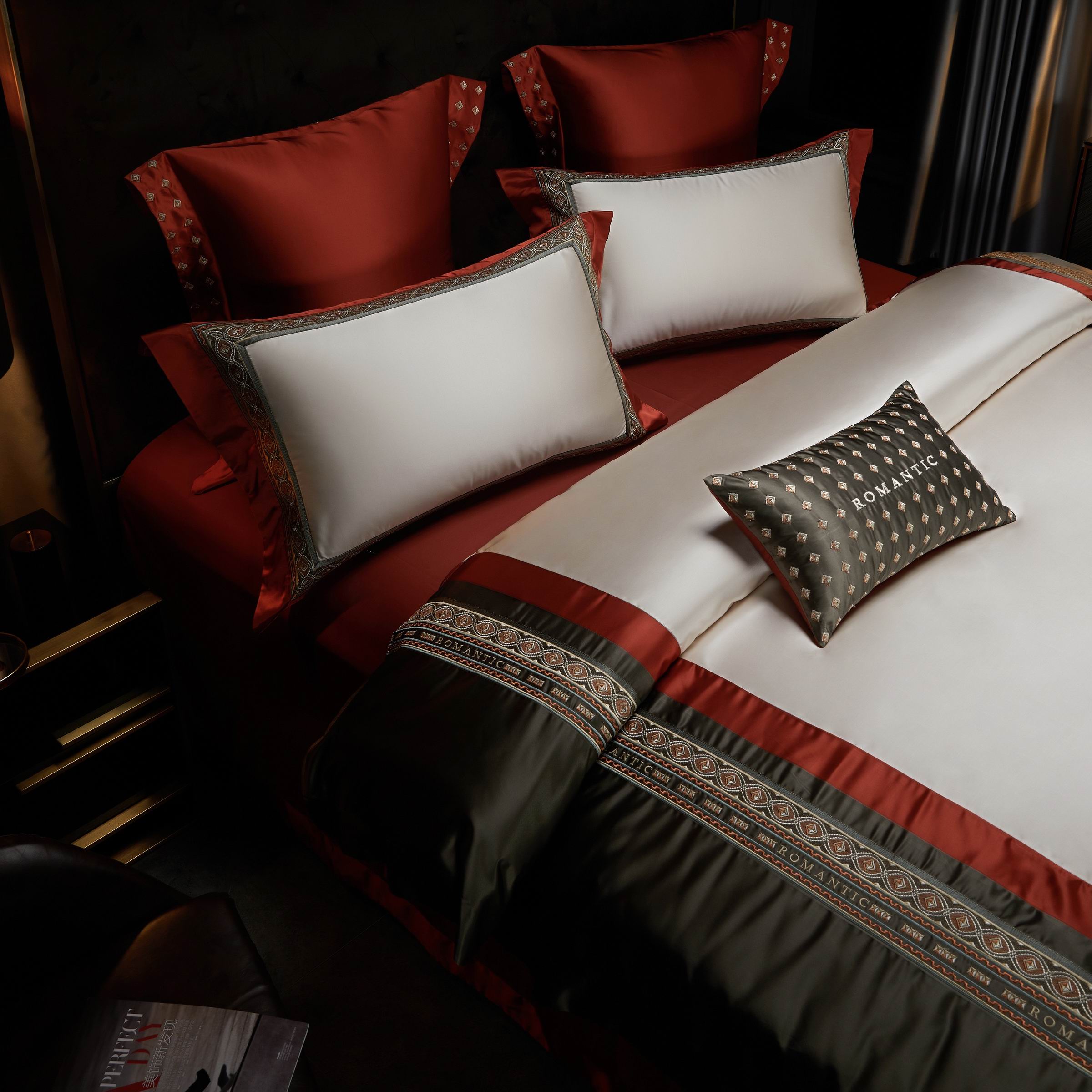 A bedding arrangement displaying a harmonious blend of ivory pillowcases bordered with crimson and accented by olive green embroidery, complemented by matching crimson pillow shams. A dark olive decorative pillow with "ROMANTIC" embroidered in white adds a modern and stylish touch. The duvet cover features alternating crimson and olive green panels, embellished with detailed embroidery for a luxurious and cohesive look. Soft lighting and a rich color palette create a warm and inviting bedroom ambiance.