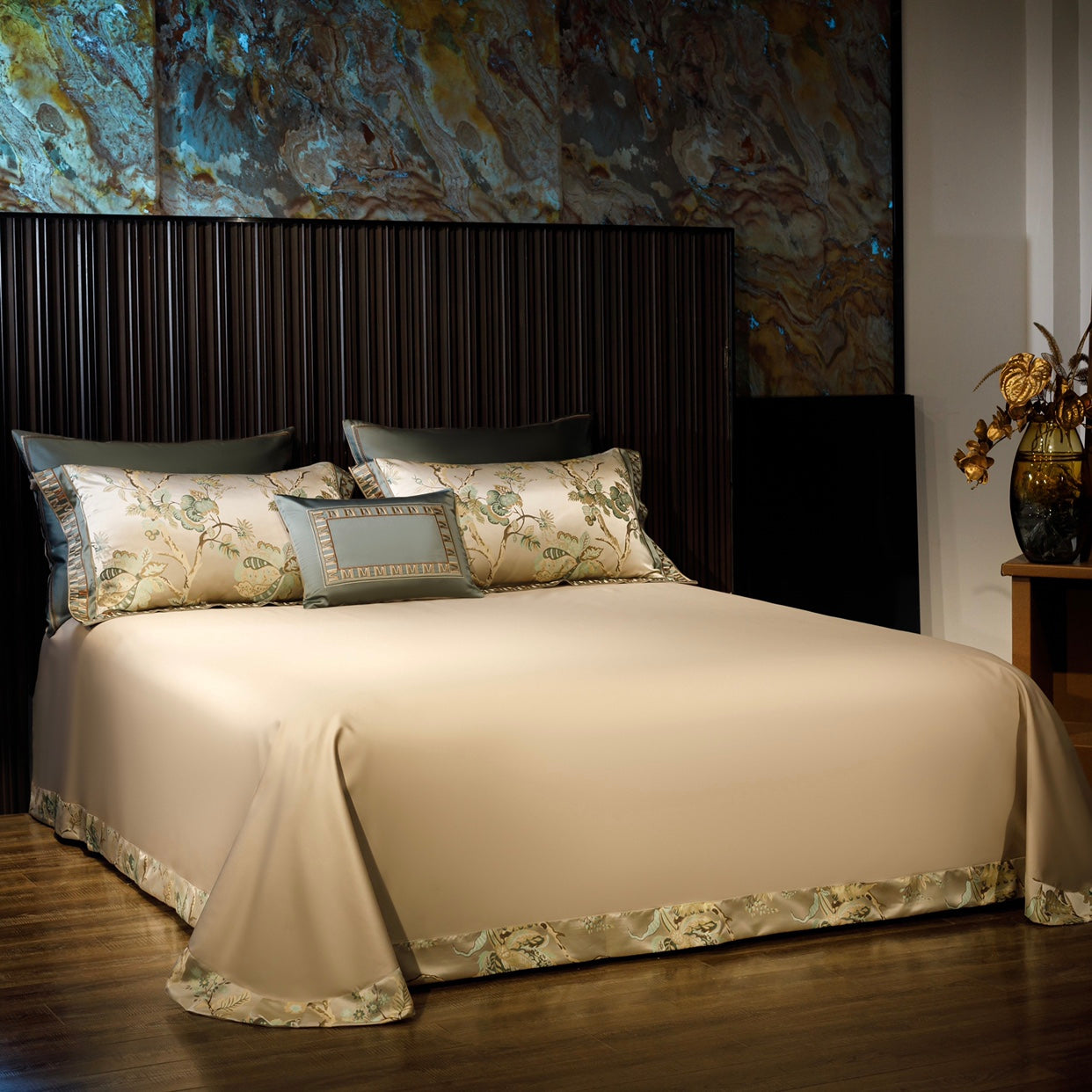 This image showcases a refined and luxurious bedding arrangement with a beige bedspread featuring a smooth, elegant texture and a decorative floral trim in muted green and ivory tones around the edges. The bed is accented with matching pillowcases adorned with delicate botanical embroidery, alongside a coordinating decorative cushion with a geometric border design. The dark wood, vertical-paneled headboard creates a striking contrast, while the marble-inspired wall panel in earthy tones adds depth and warmt