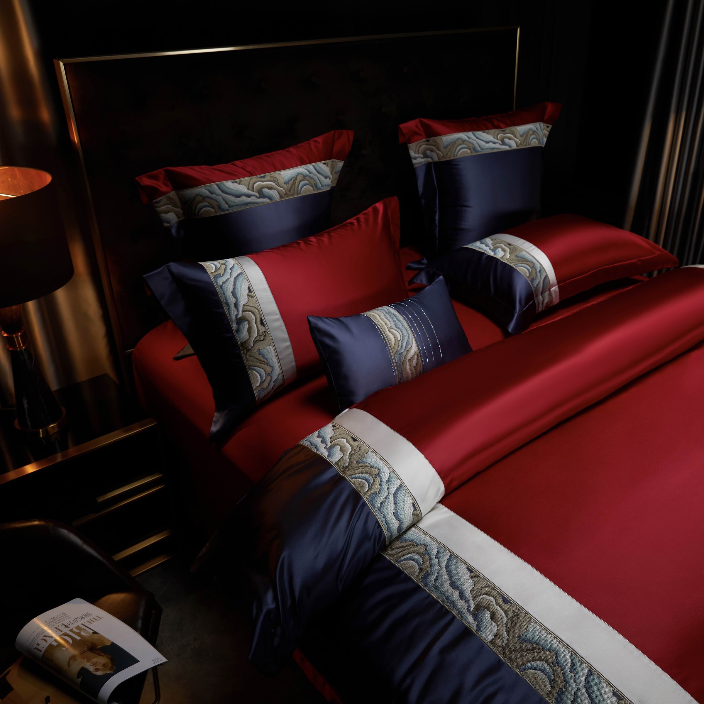 The image displays a luxurious bedding ensemble in rich burgundy and navy tones. The design is accentuated by a striking marble-patterned trim bordered in ivory, which creates a visually captivating contrast. Multiple pillow sizes, including decorative and sleeping pillows, are artfully arranged, showcasing the intricate details and satin texture. The ambient lighting and dark backdrop further emphasize the opulence and elegance of the bedding set, making it a centerpiece for a sophisticated bedroom decor.