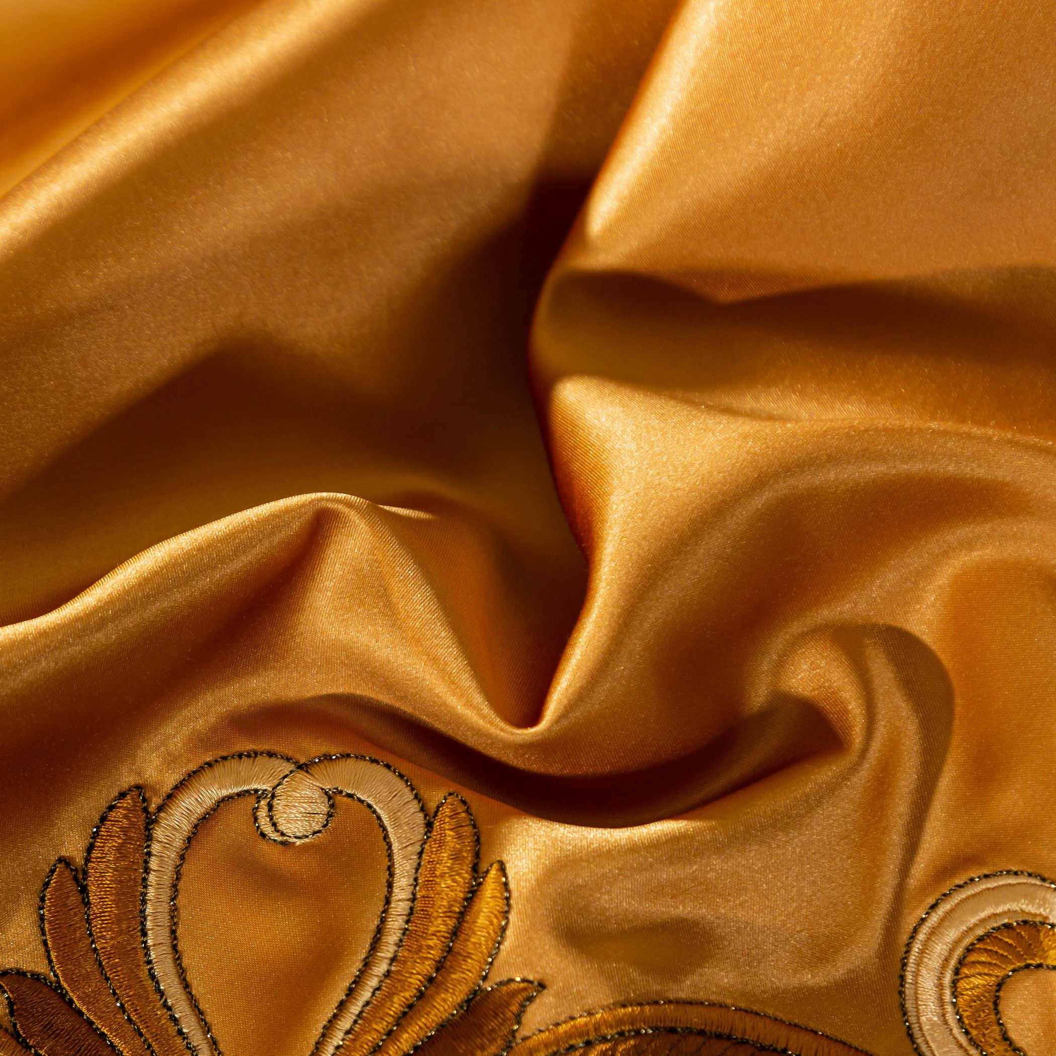 A close-up of luxurious golden-yellow satin fabric showcasing its smooth and glossy texture. The material is elegantly gathered, creating soft folds that reflect light beautifully, emphasizing its silky sheen. Visible in the corner is intricate embroidery featuring baroque-inspired scrollwork in darker gold and black threads, adding an ornate touch of sophistication to the fabric.