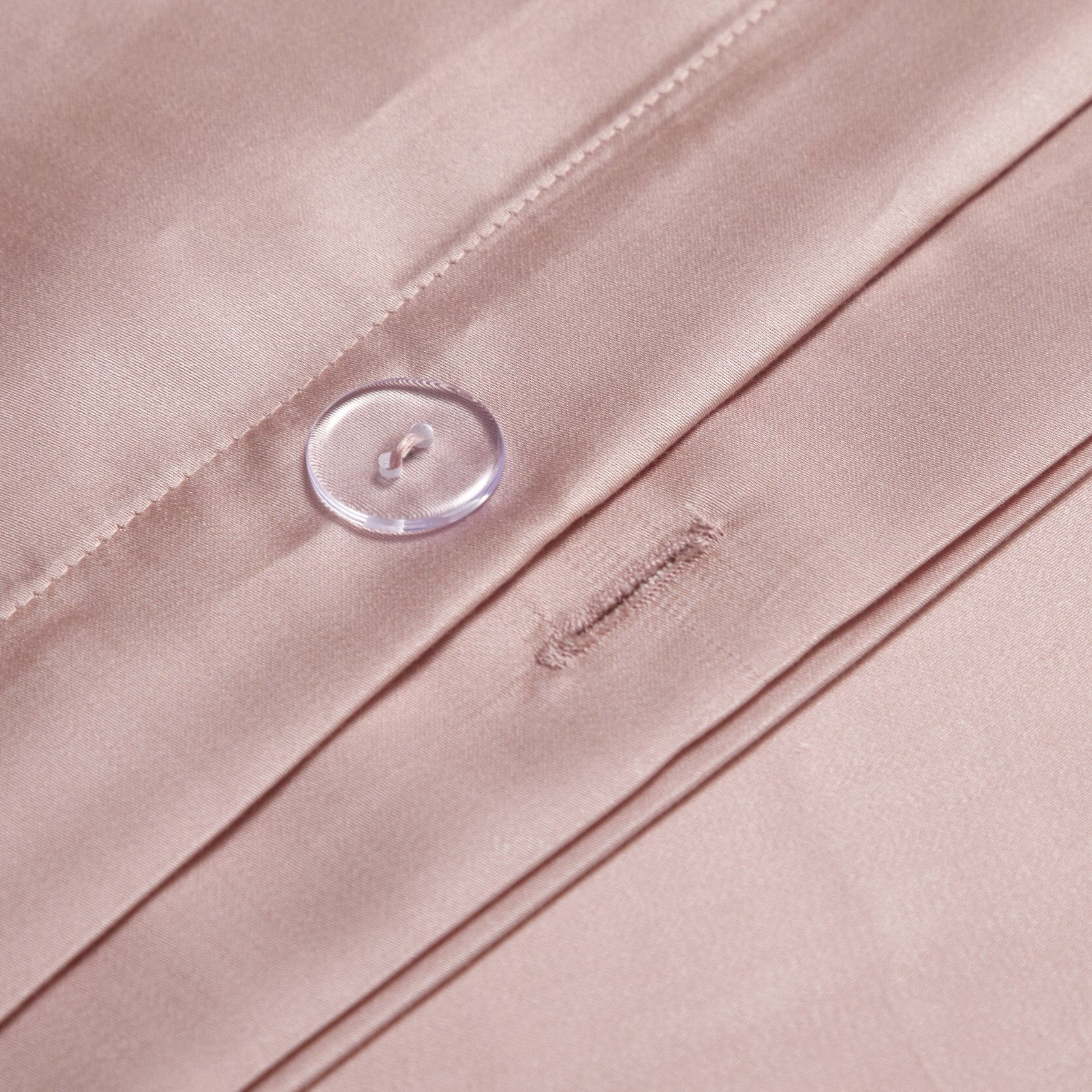 A close-up of a blush pink silk fabric with a clear button detail, showcasing precise stitching and a tailored buttonhole. The material's glossy texture highlights its luxurious quality, while the button adds a functional and aesthetic touch.